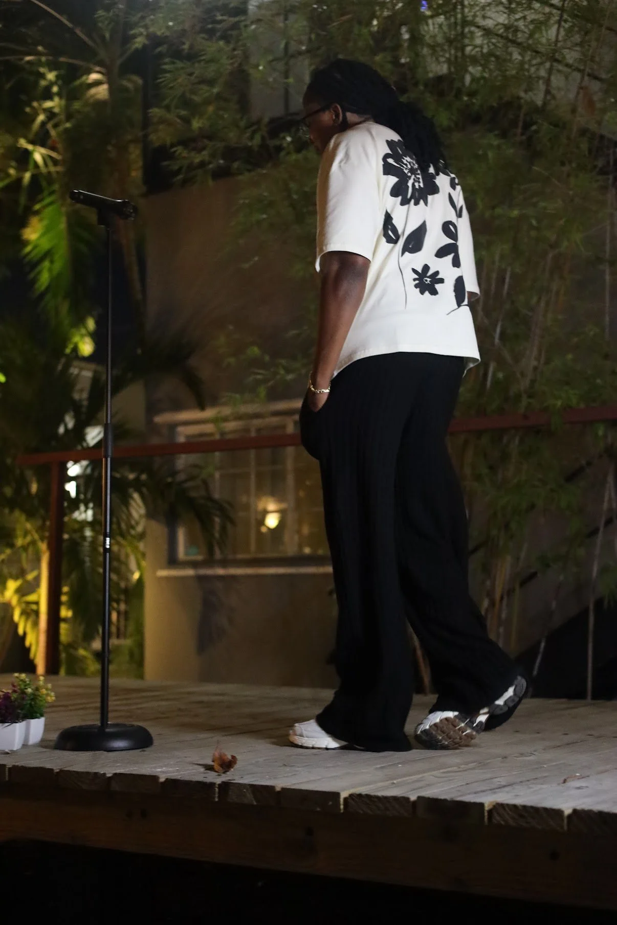 Person on stage wearing a white shirt with floral design, black pants, and walking on a wooden platform at night.