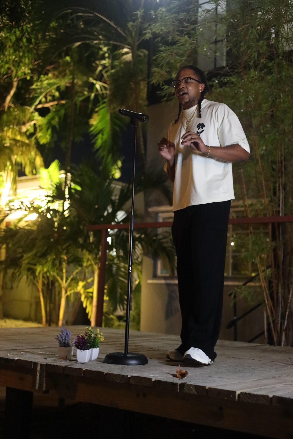 A person with long braided hair and glasses standing on a wooden stage, singing or speaking into a microphone, with potted plants nearby and lush greenery in the background at night.