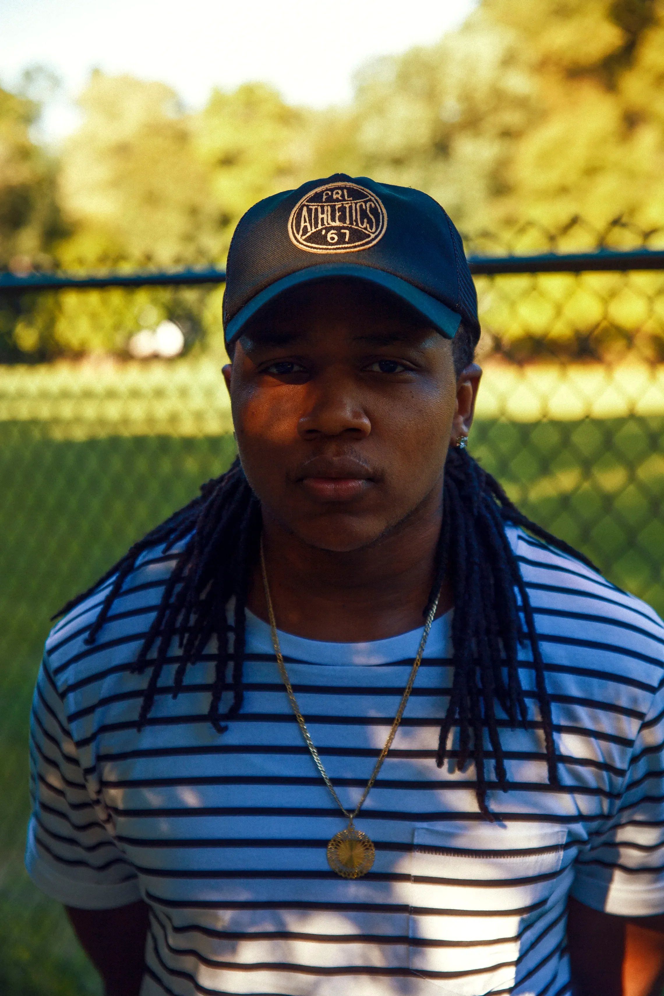 Young man with long dreadlocks wearing a striped T-shirt, a gold chain with a pendant, and a baseball cap, standing outdoors near a fence with trees in the background.