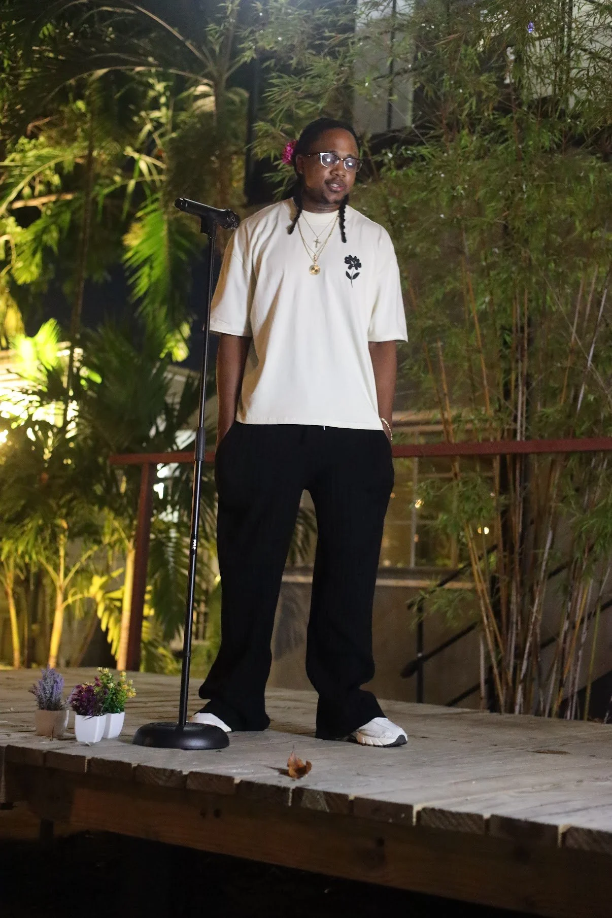 A person with glasses and braided hair tied with a pink flower, wearing a beige T-shirt and black pants, stands on a wooden stage with a microphone. They are outdoors with lush green plants in the background.