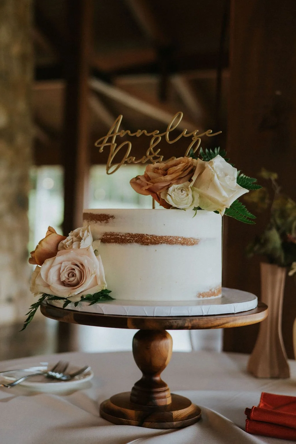 Wedding cake decorated with fresh roses and greenery, with a gold topper inscribed with the names "Annaleigh and Daniel".