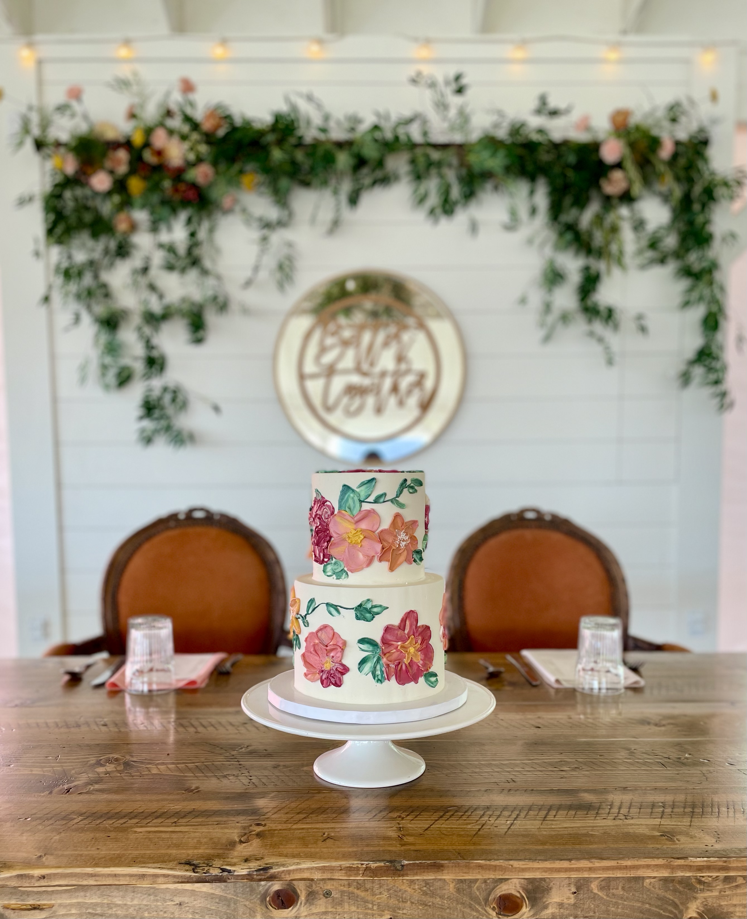 A three-tiered wedding cake decorated with pink and orange flowers and green leaves, placed on a white cake stand on a wooden table, with chairs, glasses, and napkins behind it, in a decorated indoor setting.