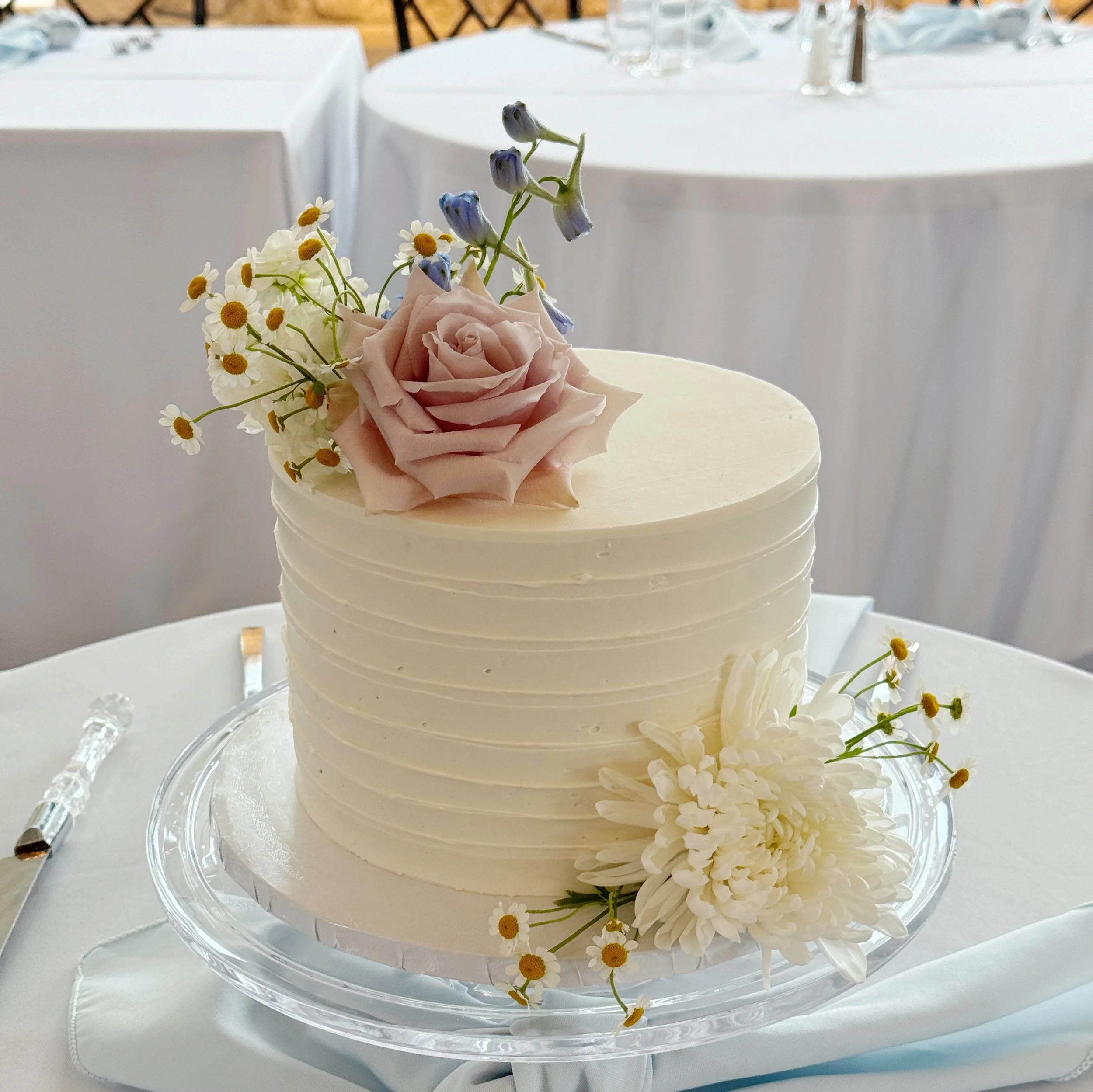 A white, textured wedding cake topped with a pink rose, small daisies, blue flowers, and a white chrysanthemum, sitting on a glass cake stand on a white tablecloth.