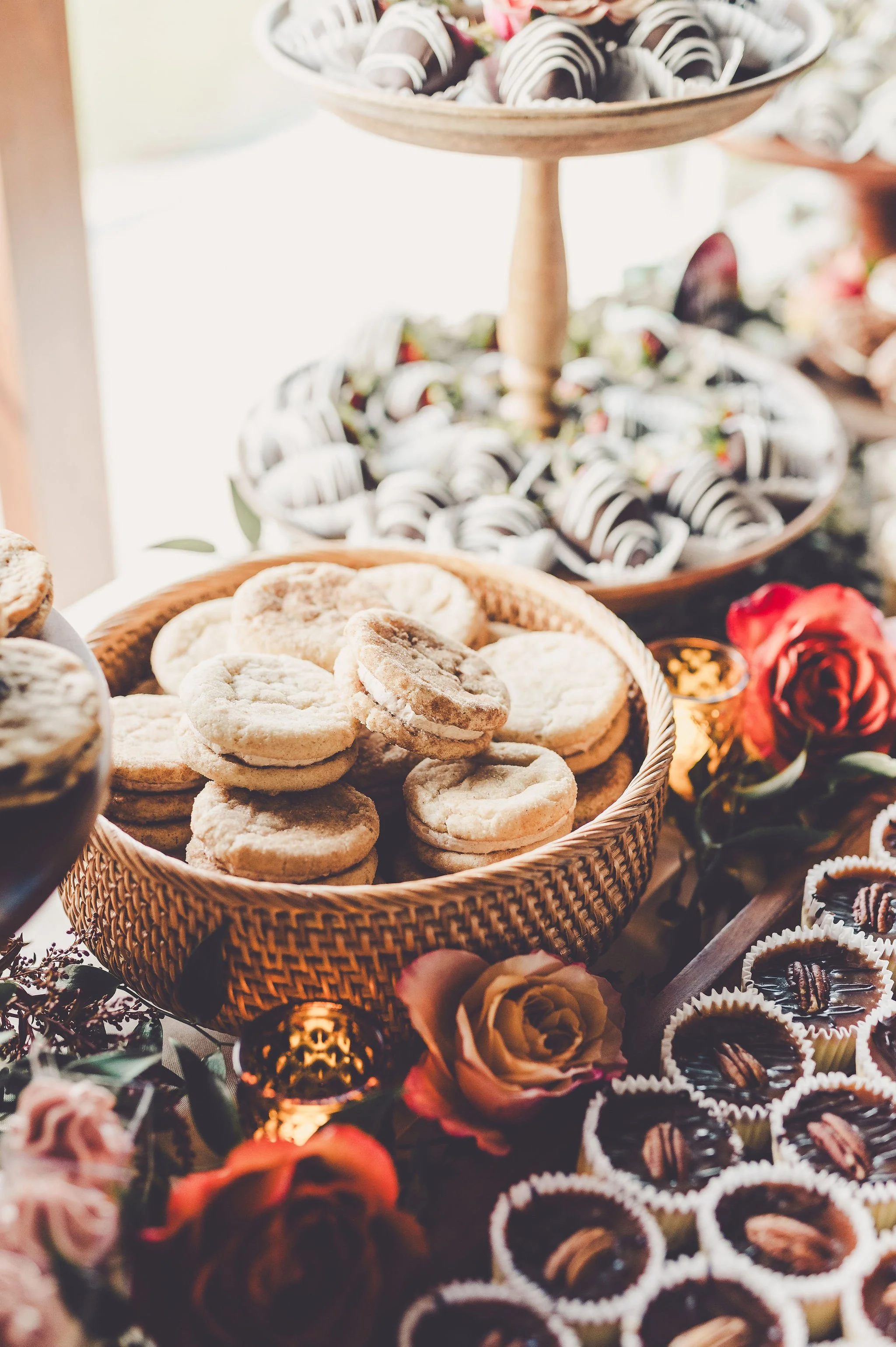 A display of assorted cookies and chocolates, with some decorated with icing and others with pecans, arranged on a table decorated with roses and small decorative candle holders.