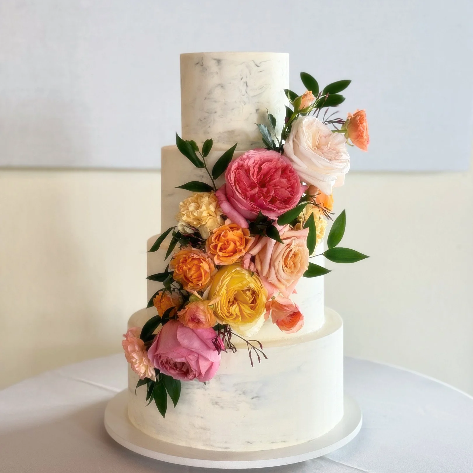 A tiered white marble wedding cake decorated with a cascading arrangement of pink, peach, yellow, and cream flowers and green leaves.