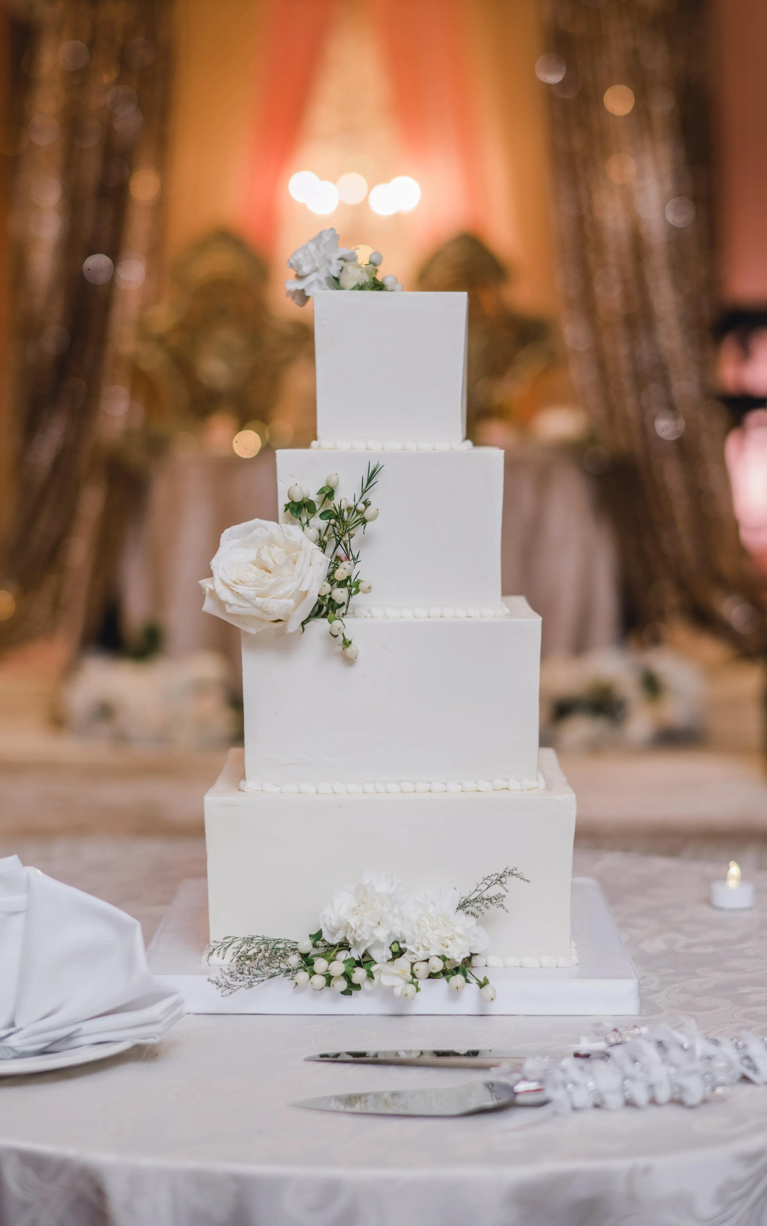 Four-tiered white wedding cake decorated with white flowers and greenery, placed on a table with cake utensils, in a decorated banquet hall.