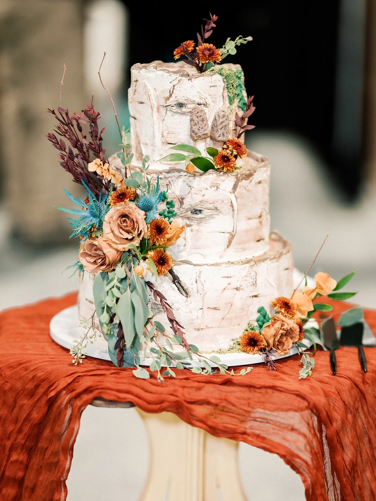 Three-tiered wedding cake with rustic bark appearance, decorated with orange and peach flowers and green foliage, on a white cake board, on a table covered with an orange cloth.