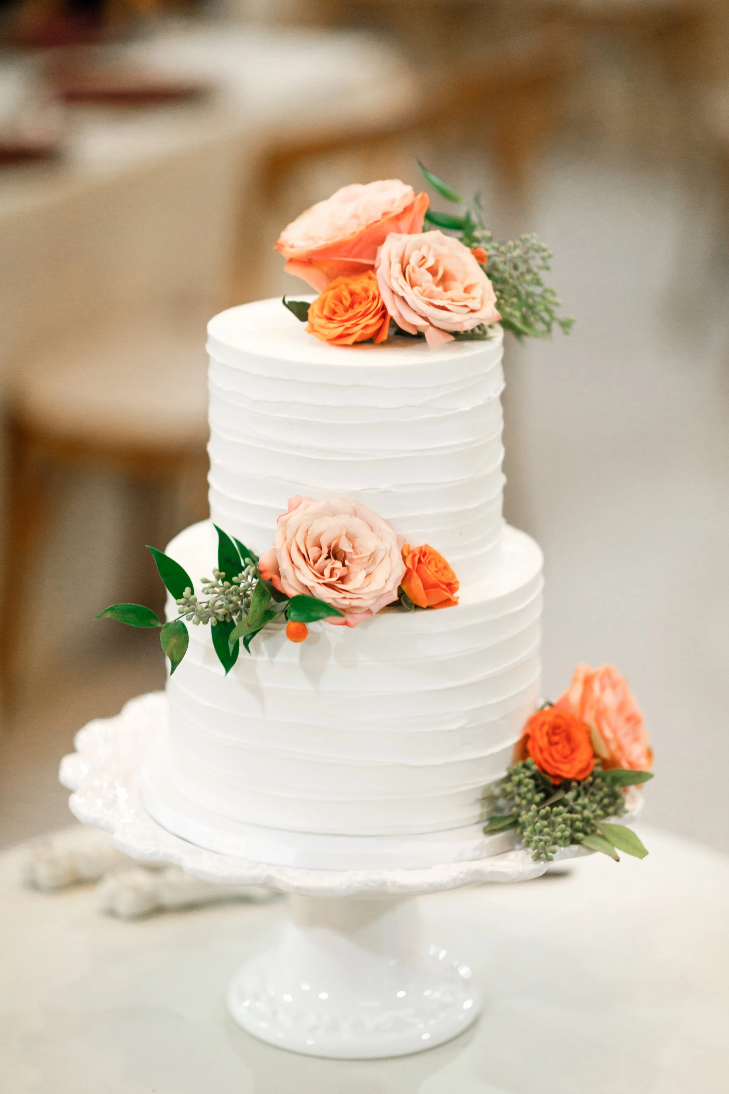 A three-tier white wedding cake decorated with pink and orange flowers and green foliage, placed on a white cake stand.