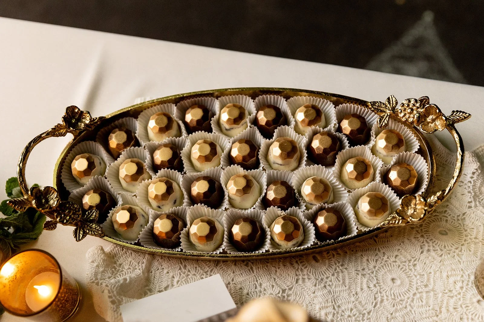 Ornate gold tray filled with white and dark chocolates decorated with gold geometric patterns, set on a white lace tablecloth with a lit candle nearby.