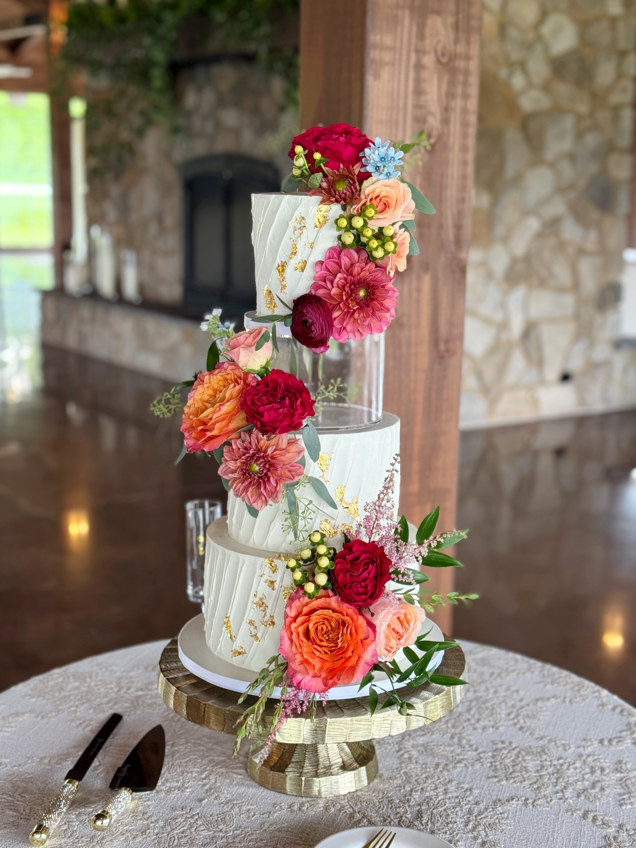 Three-tiered wedding cake decorated with colorful flowers and gold leaf accents, displayed on a wooden cake stand with a tablecloth and cake-cutting utensils nearby.