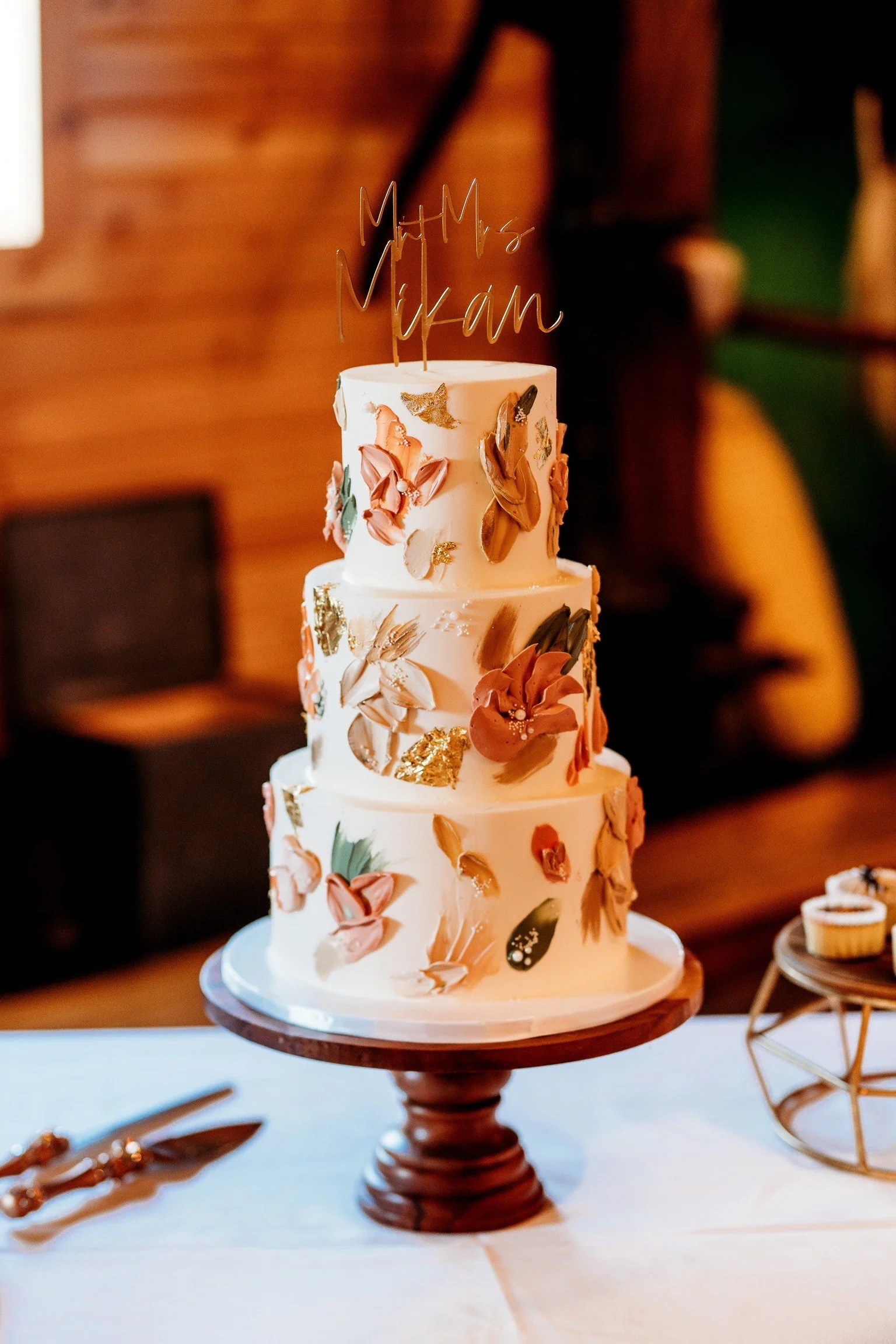 A four-tiered wedding cake with floral and gold accents on a wooden cake stand, featuring a "Mrs. & Mr." cake topper, on a decorated table in a rustic setting.