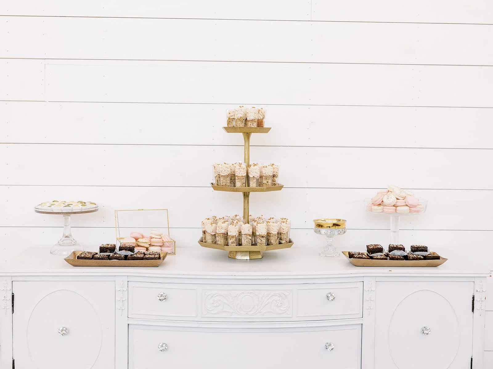 Dessert table with cupcakes, macarons, and brownies on white sideboard against white wooden wall.