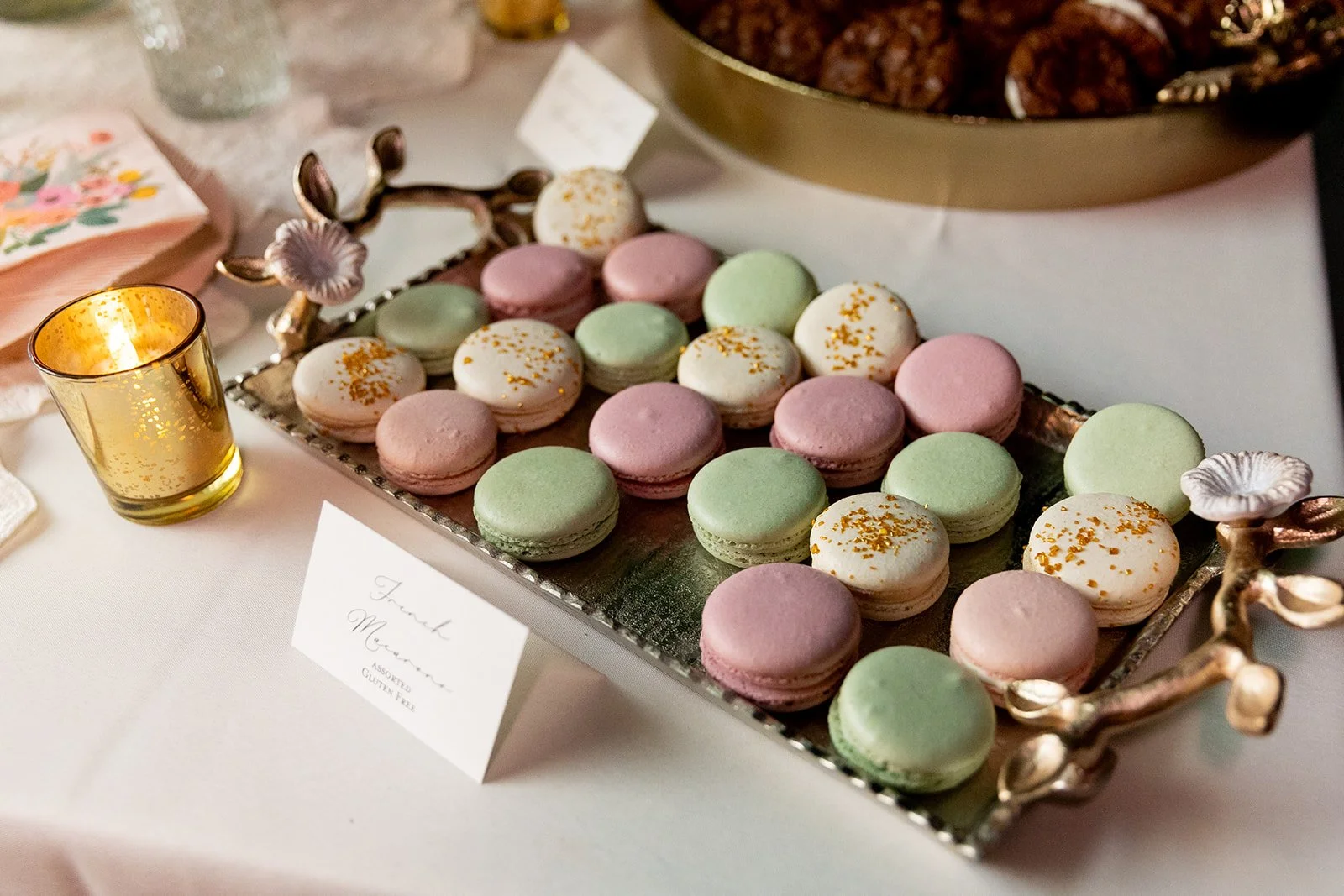 Assorted pastel-colored macarons arranged on a decorative silver tray with rose-shaped handles, next to a gold votive candle.