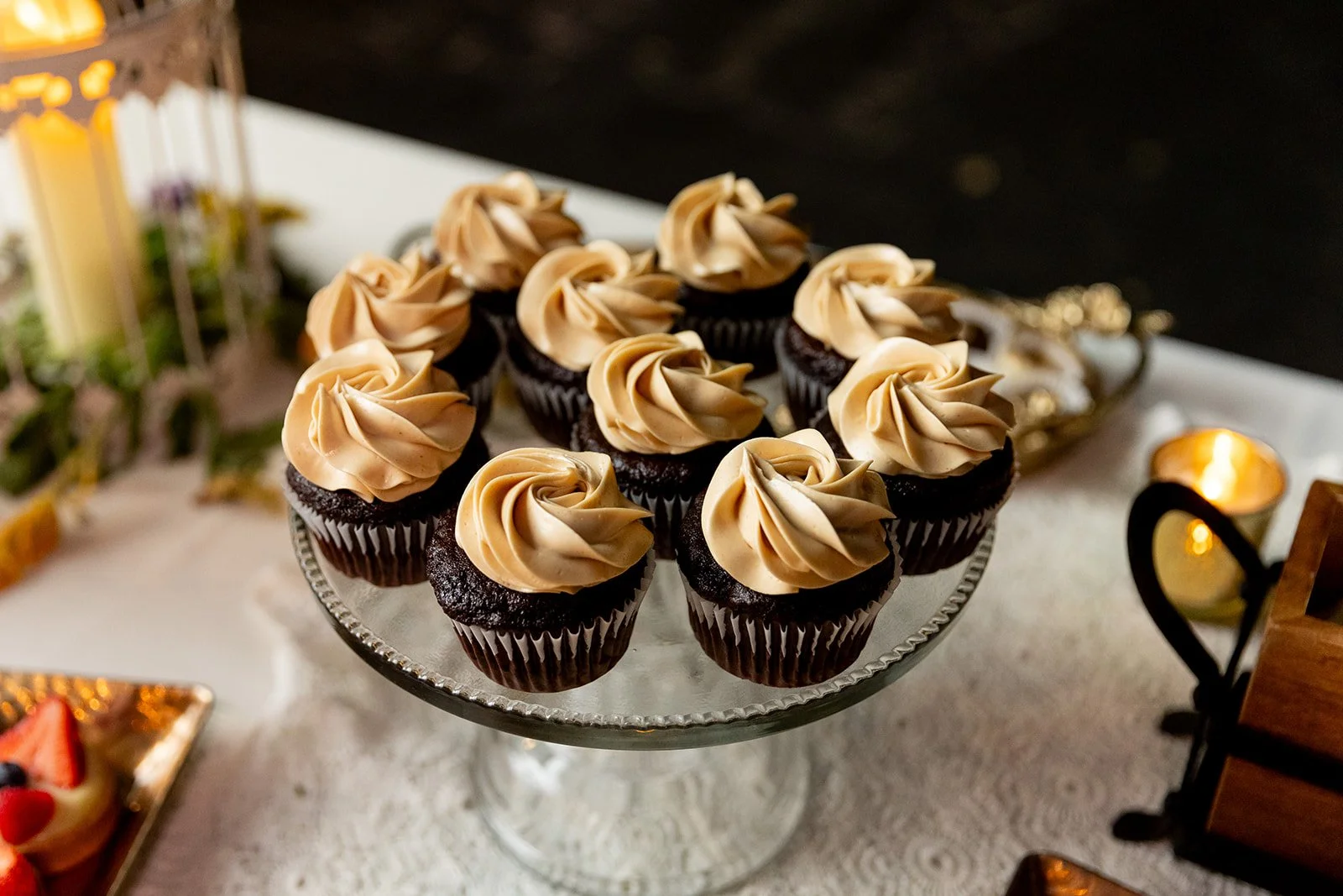 A glass cake stand holding chocolate cupcakes with beige frosting swirls.