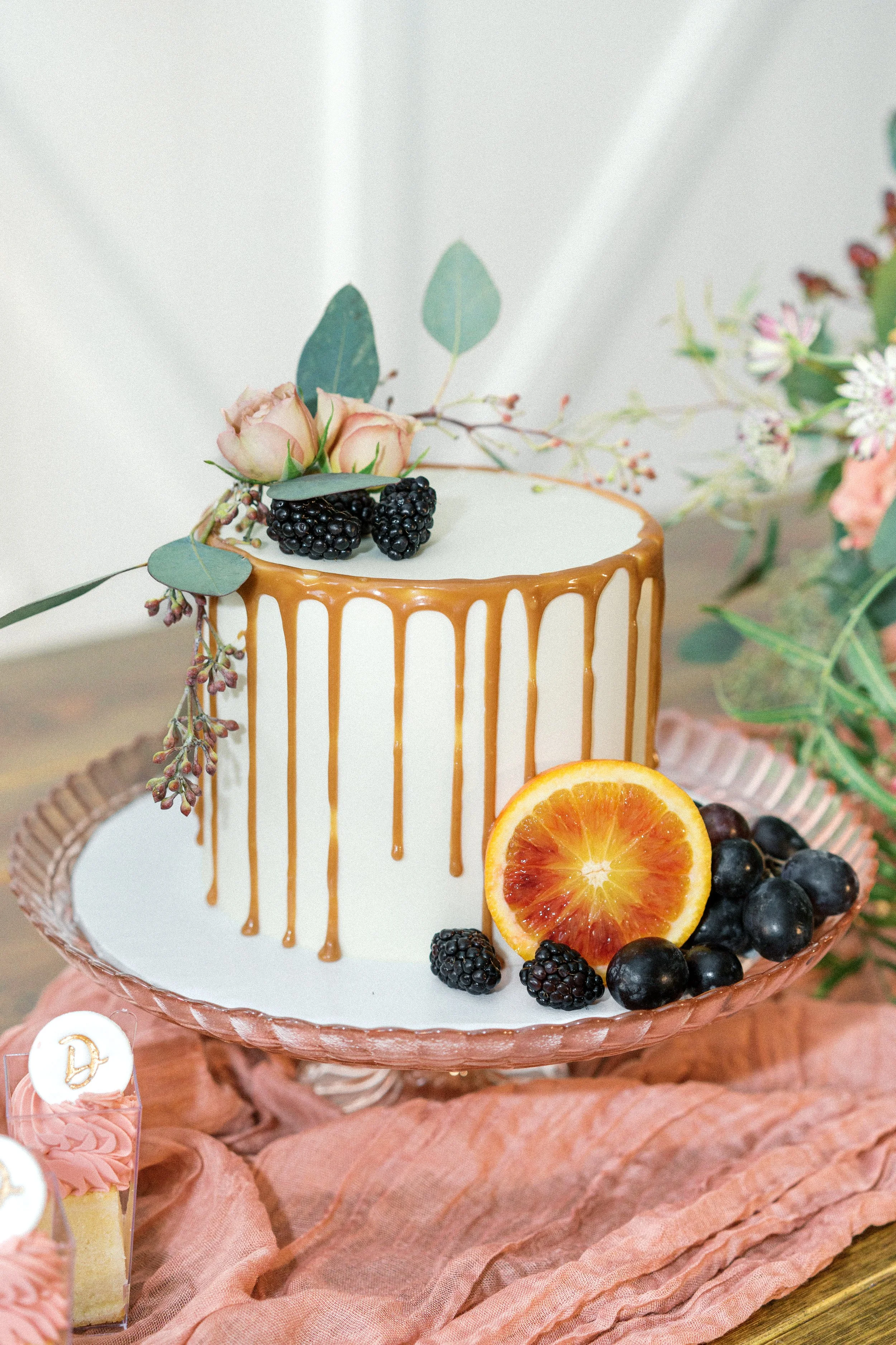 A white cake with caramel drip decoration, adorned with pink roses, blackberries, and an orange slice, placed on a pink glass cake stand surrounded by flowers.