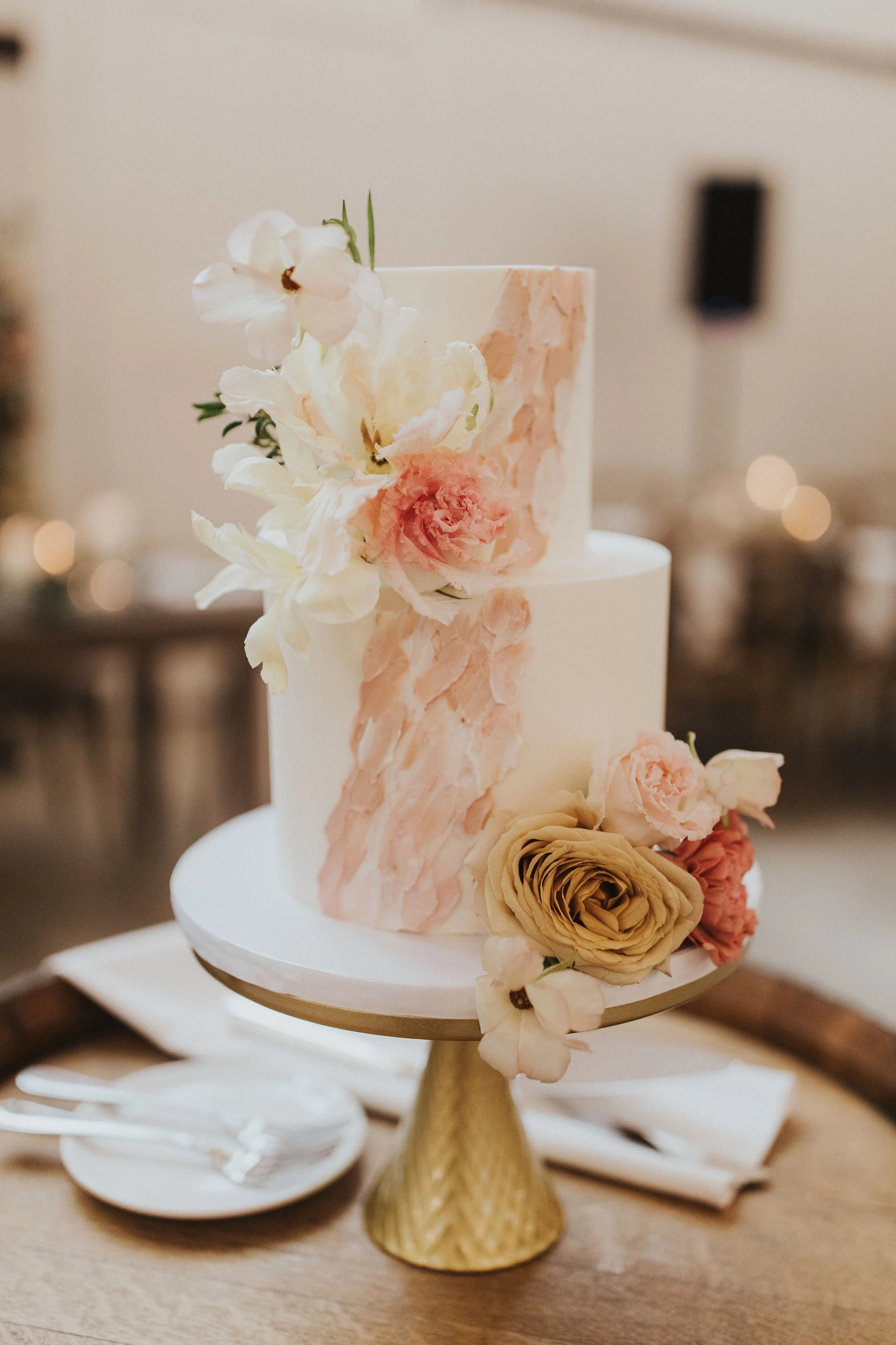 A two-tiered wedding cake decorated with fresh flowers, placed on a gold cake stand.