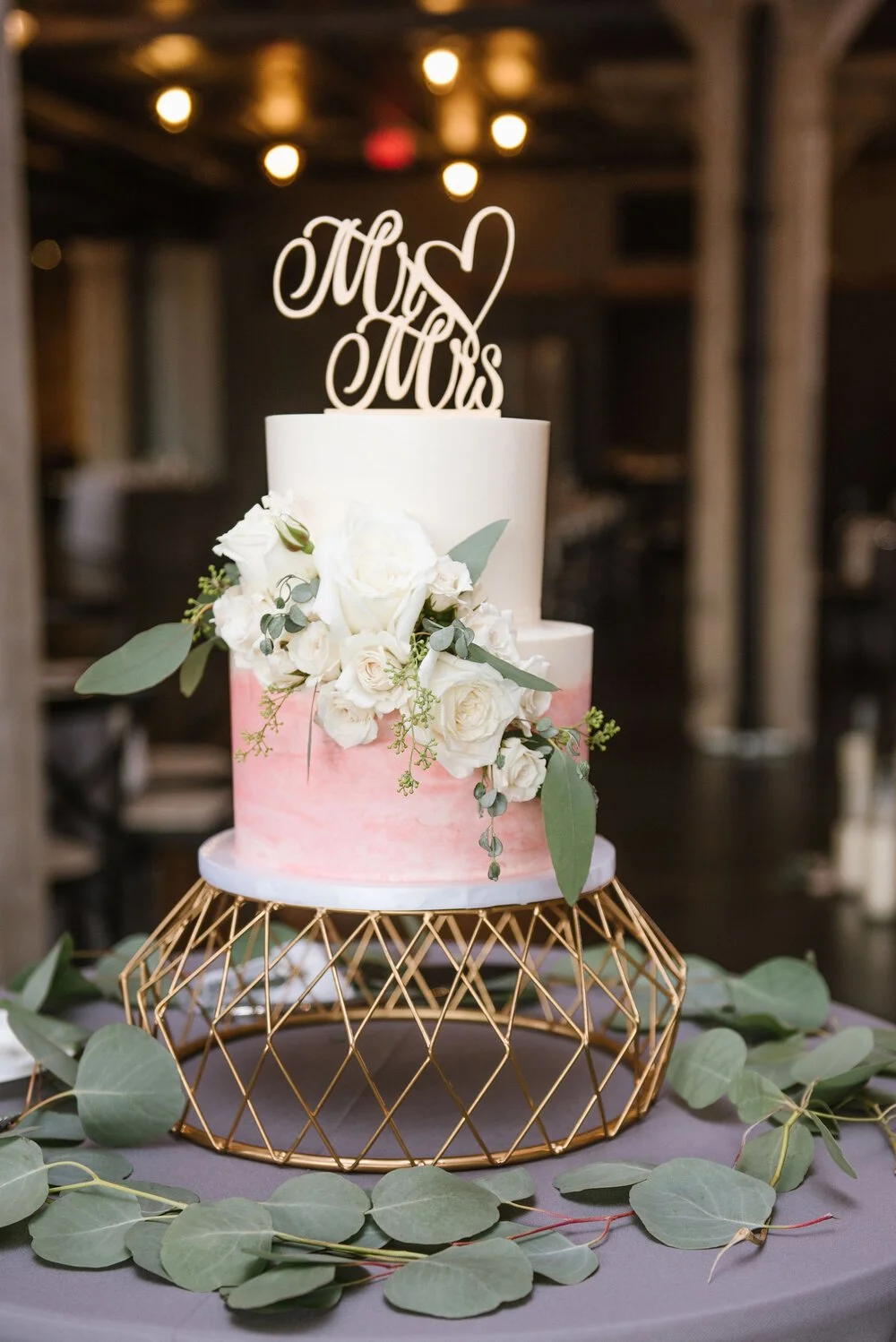 A two-tier wedding cake decorated with white roses and eucalyptus, topped with a "Mrs & Mrs" cake topper, on a gold geometric stand surrounded by eucalyptus leaves.