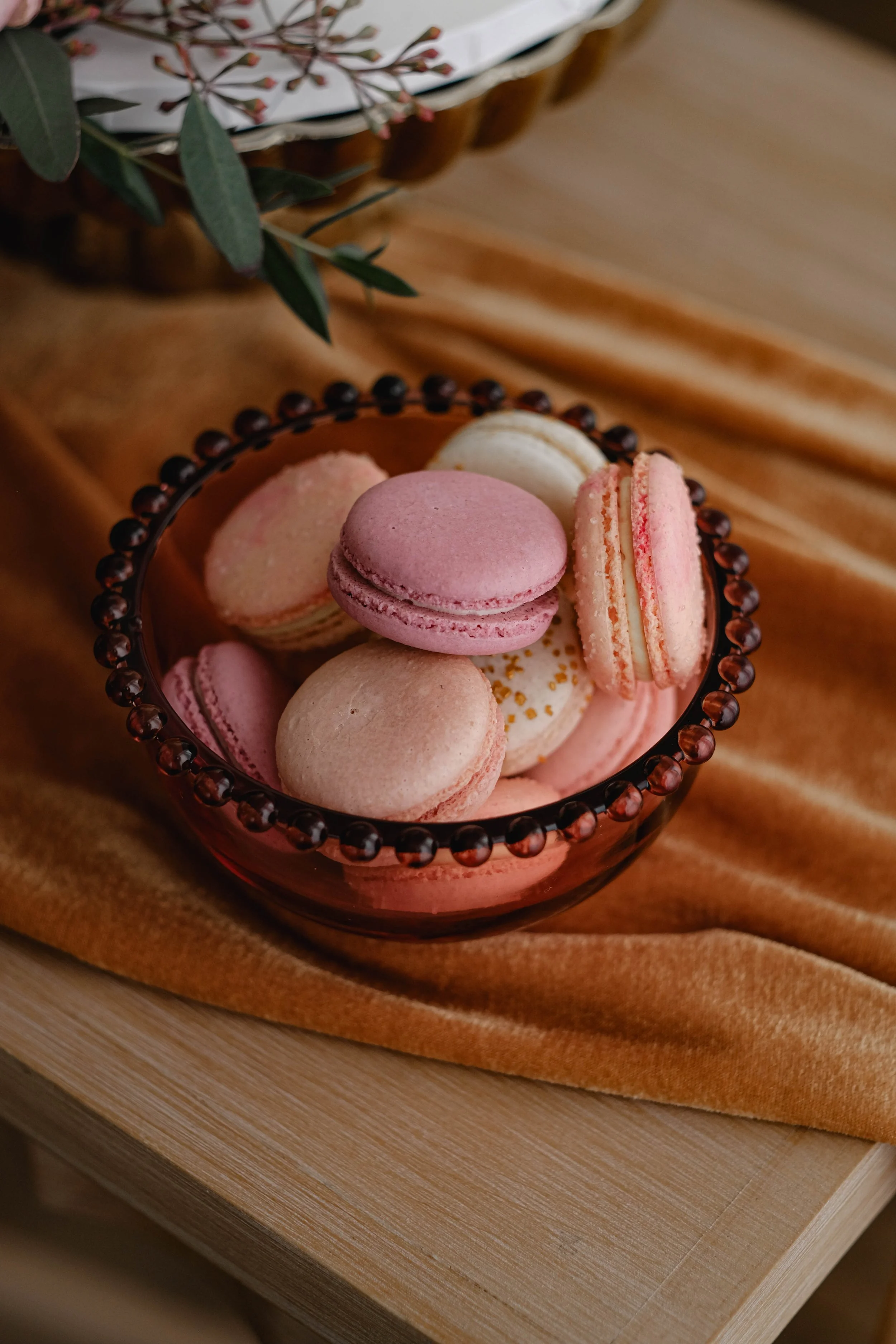 A bowl of pink and white macarons on a wooden surface, with a golden brown velvet cloth underneath and a decorative plant nearby.