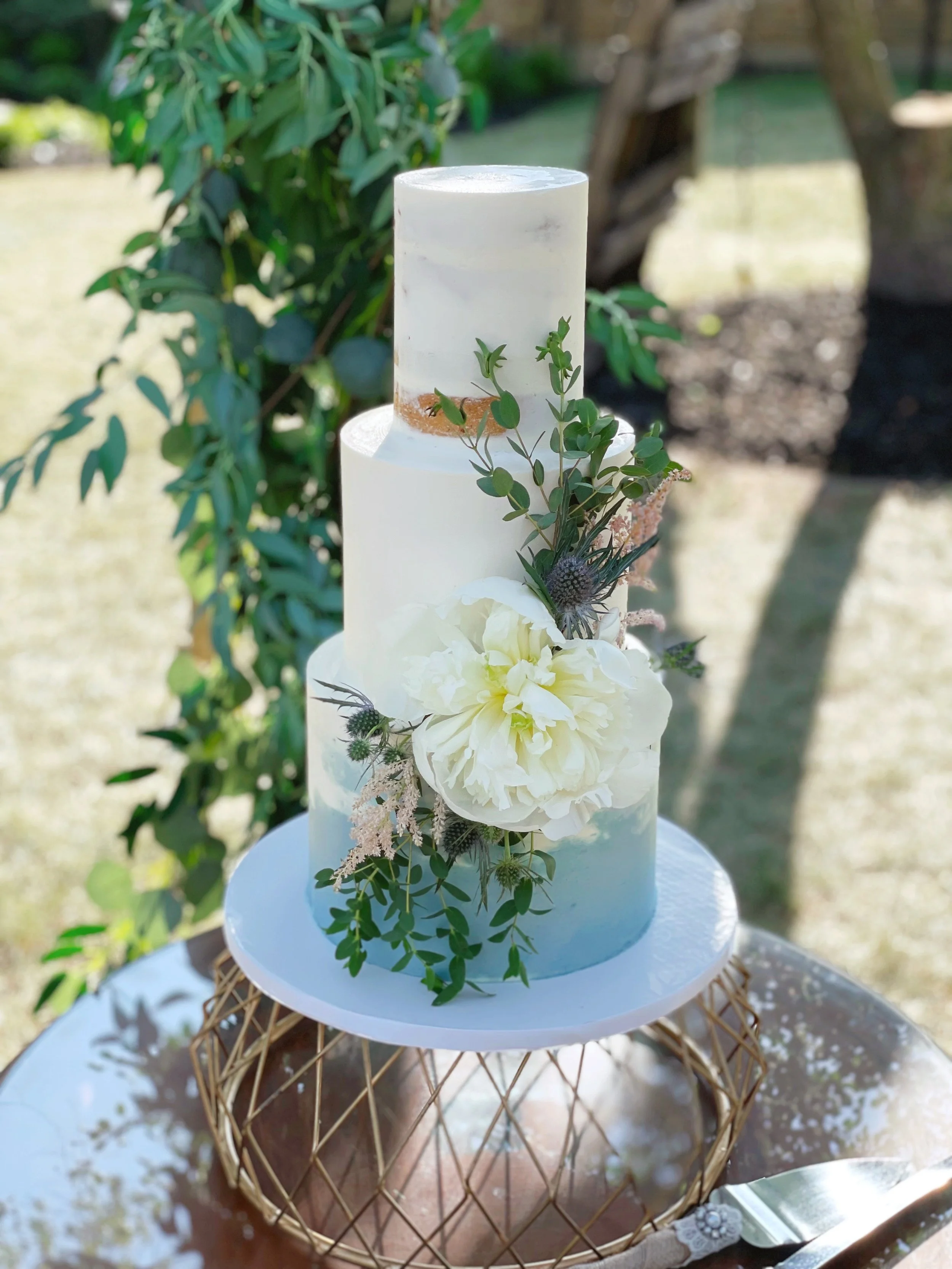 A three-tiered white wedding cake decorated with white flowers and greenery, placed outdoors on a reflective metallic surface.