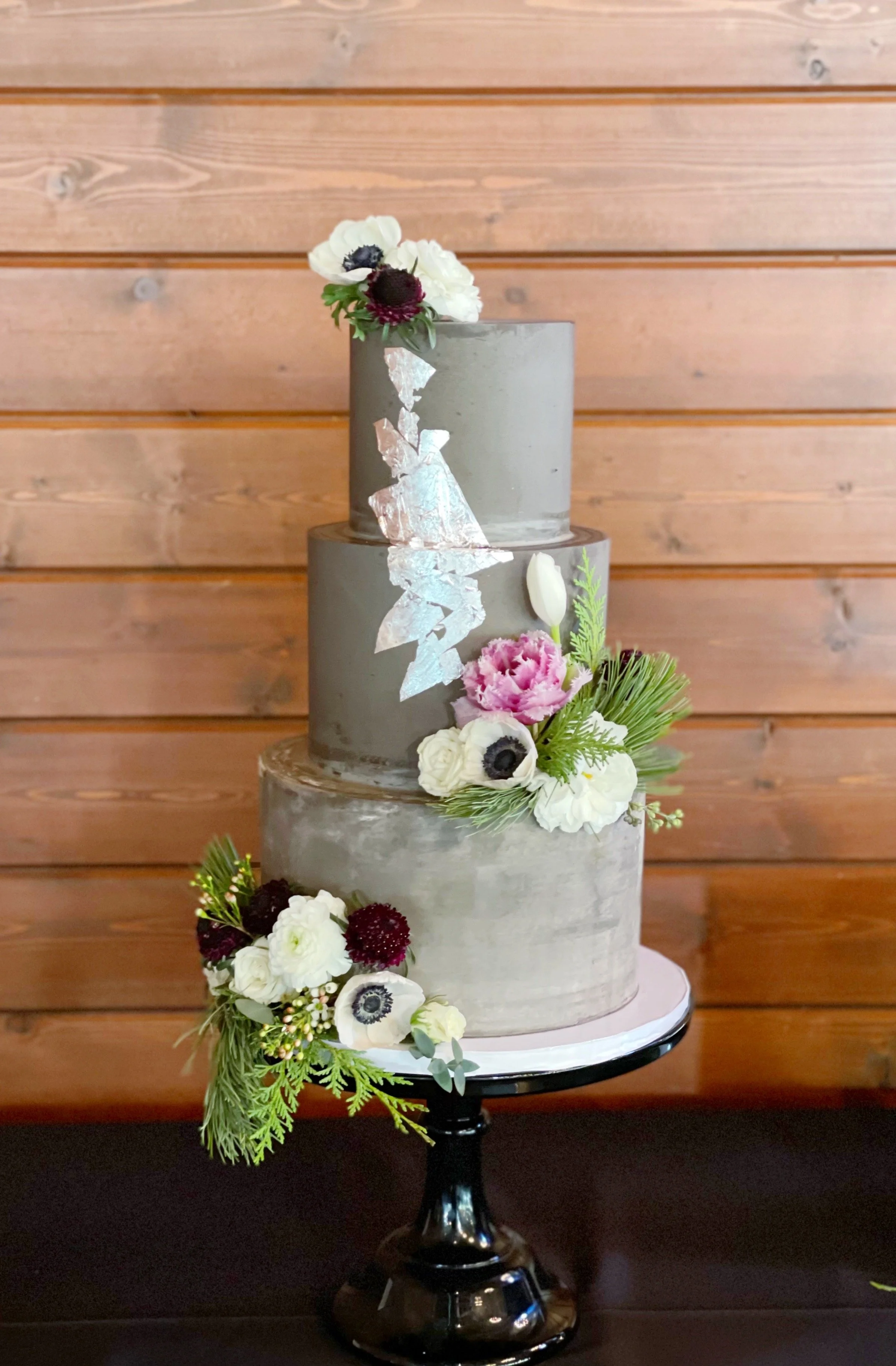 A three-tiered gray wedding cake decorated with fresh flowers and greenery, placed on a black cake stand against a wooden wall background.