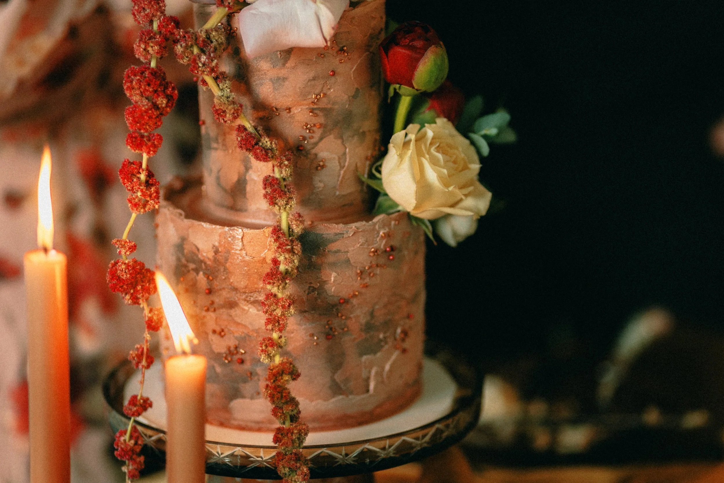A two-tiered cake decorated with fresh flowers, including white roses and red buds, with red and green foliage. The cake has a marbled or textured icing in earth tones and is displayed on a decorative stand with a black and gold pattern. There are two lit candles with orange flames casting a warm glow nearby.