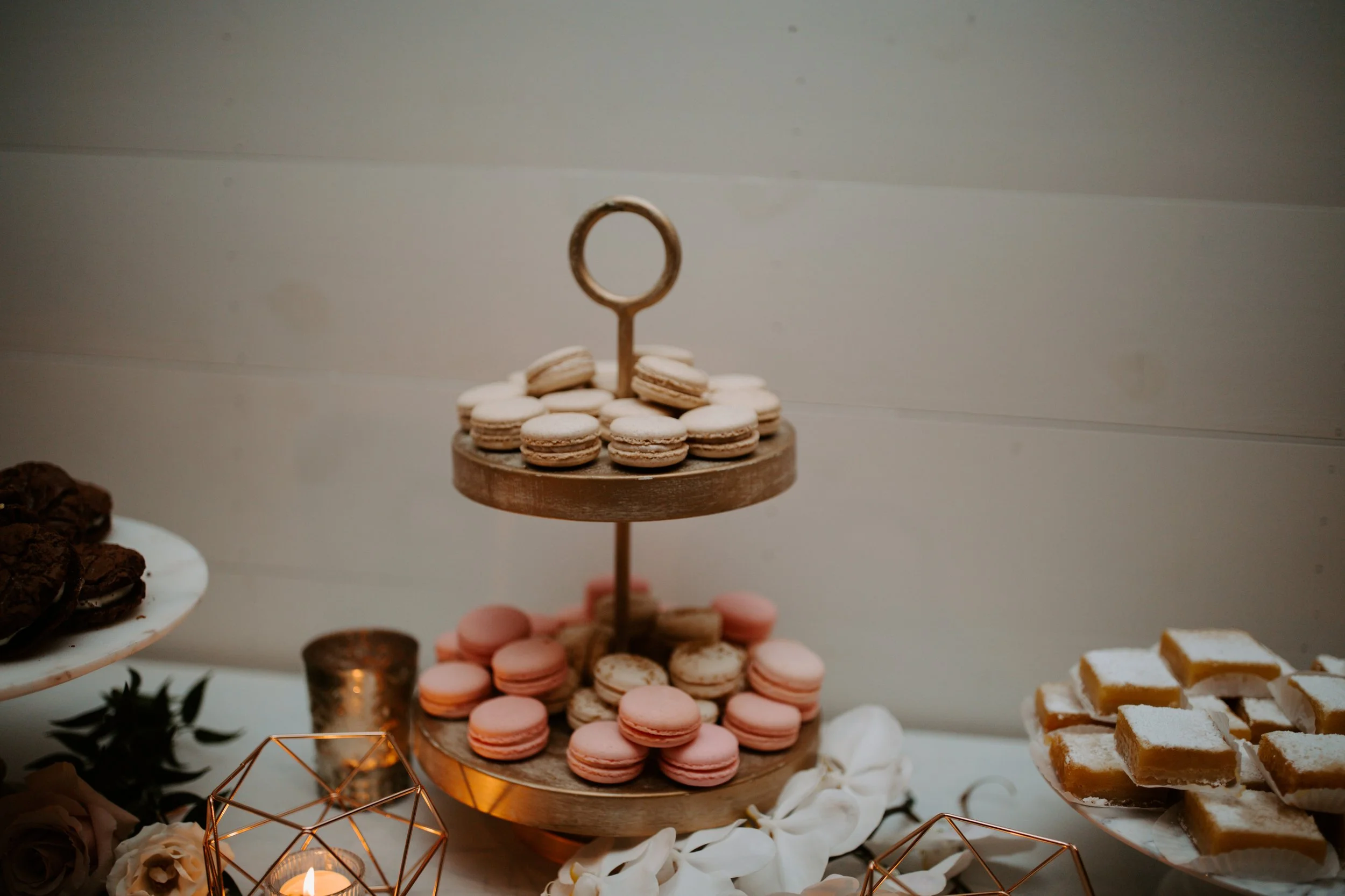 A dessert table with a two-tiered stand of macarons, pink and white, topped with a ring-shaped handle, surrounded by other desserts, candles, and decorative flowers.