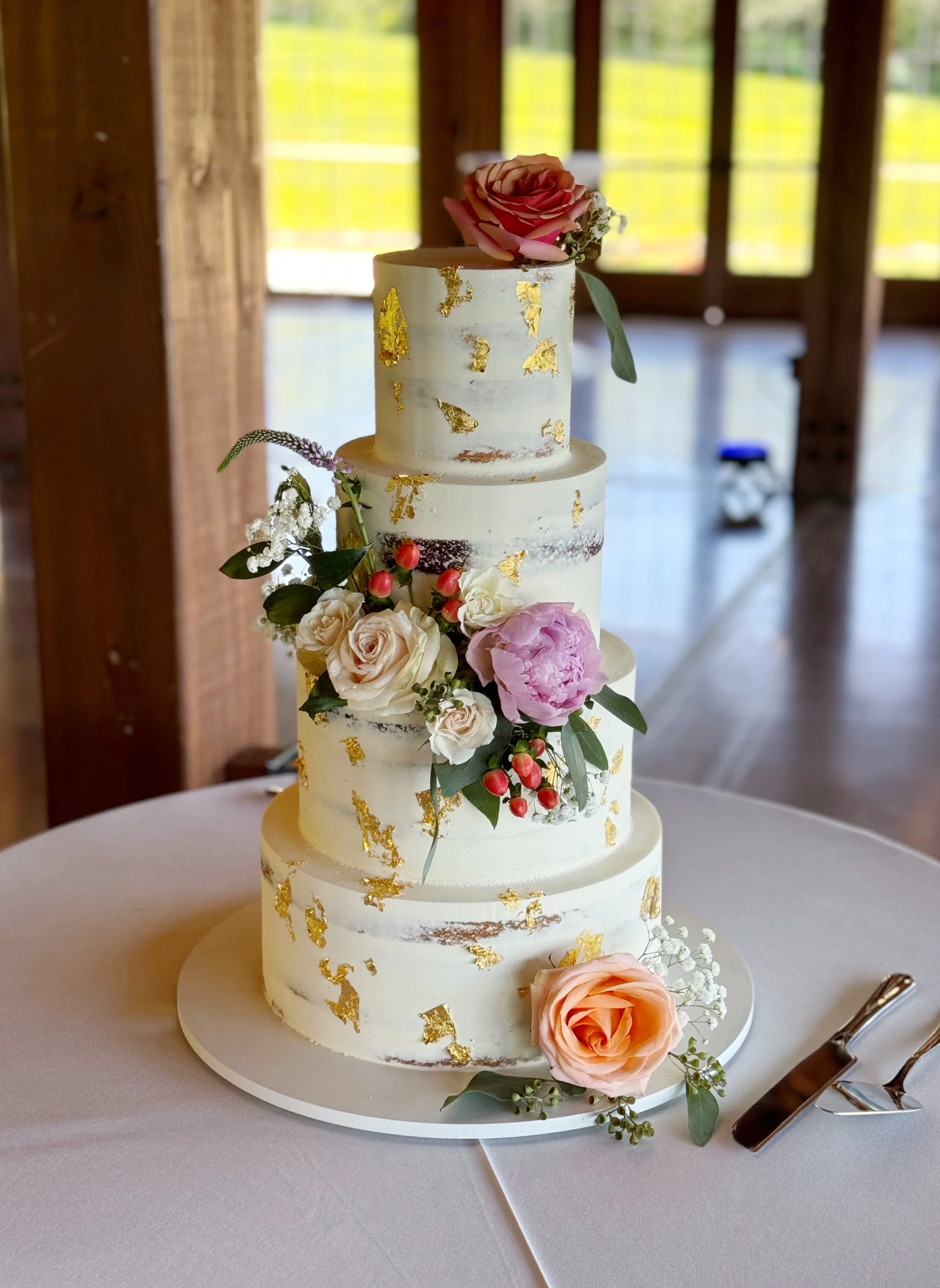 A four-tier wedding cake decorated with fresh flowers and gold leaf, placed on a round table with a knife and cake server beside it.