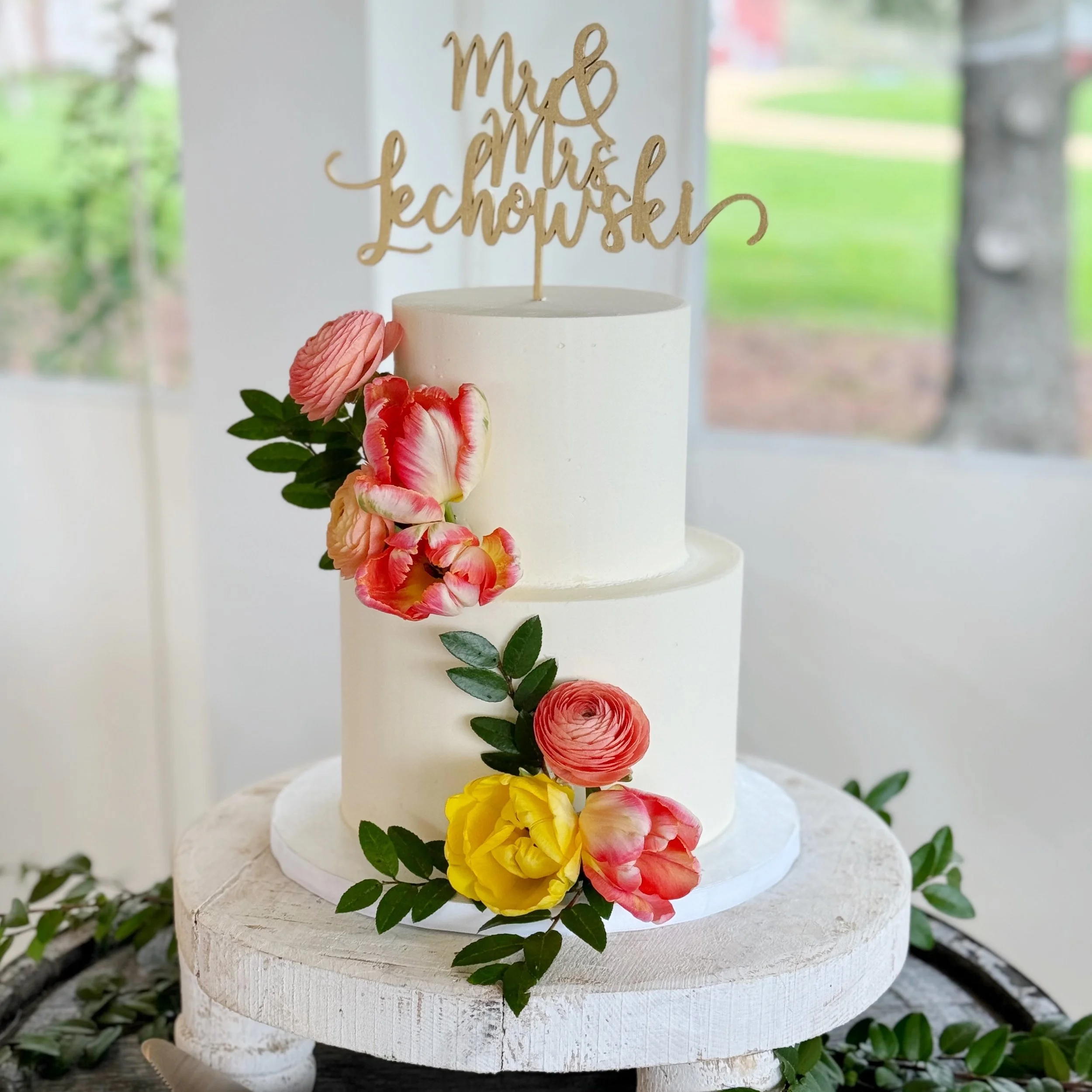 A two-tier white wedding cake decorated with pink, yellow, and orange flowers and green leaves. A gold topper reads 'Mr & Mrs Lechowski.' The cake is on a round white wooden stand.