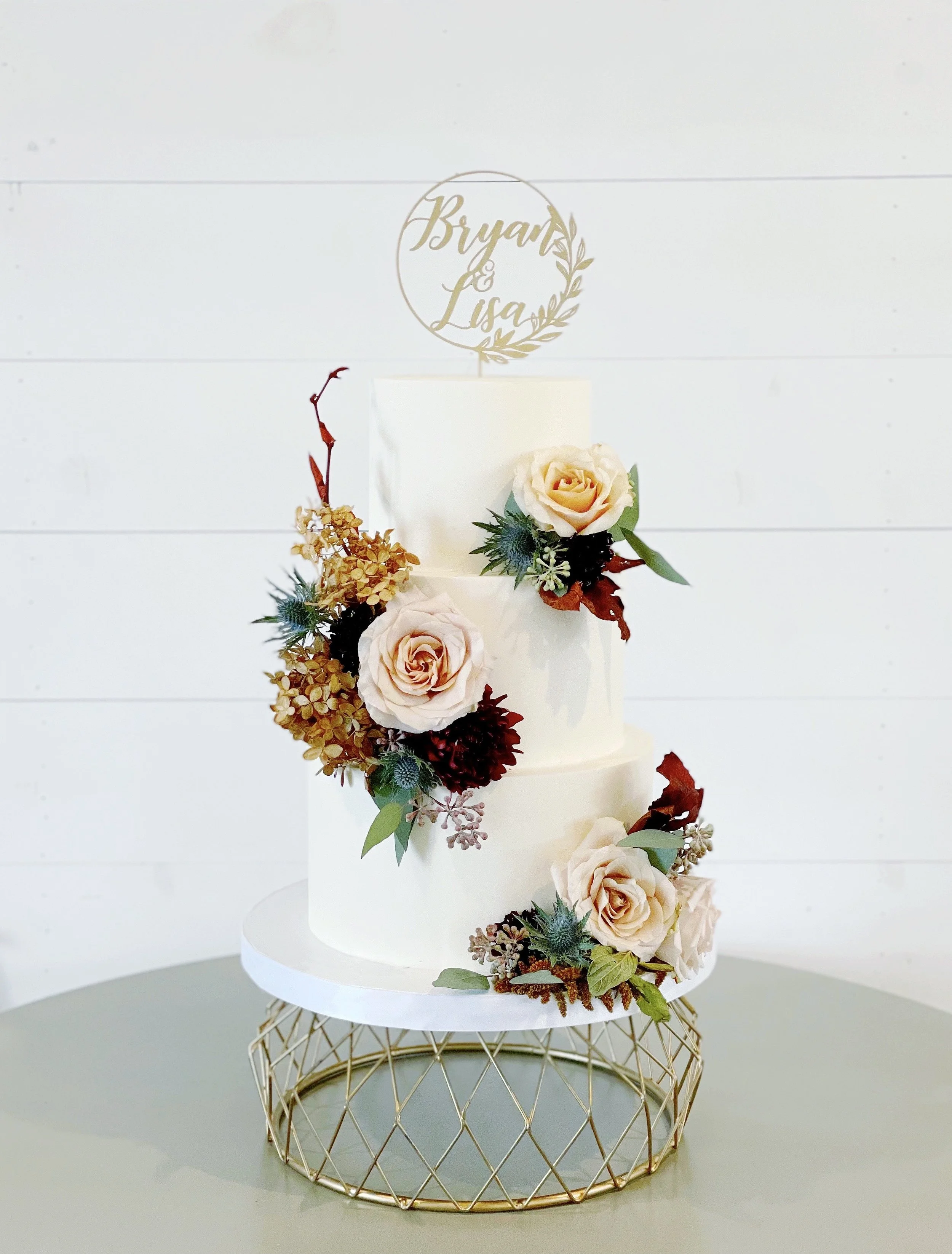 A three-tier white wedding cake decorated with cream-colored roses, greenery, and dried flowers, placed on a gold wire stand with a circular base.