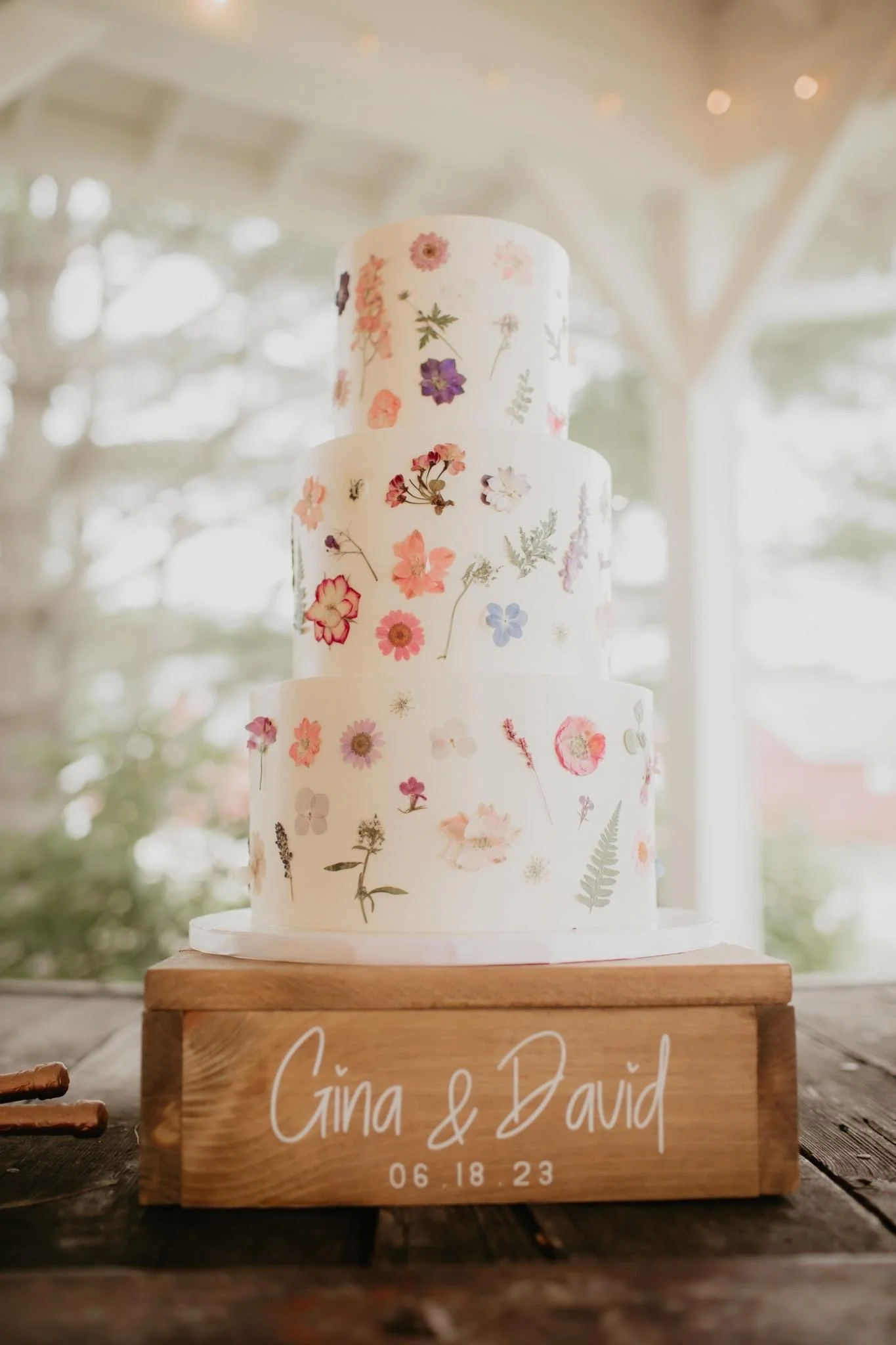 A three-tiered wedding cake decorated with pressed flowers and mounted on a wooden stand with the names 'Gina & David' and the date '06.18.23' written on it.