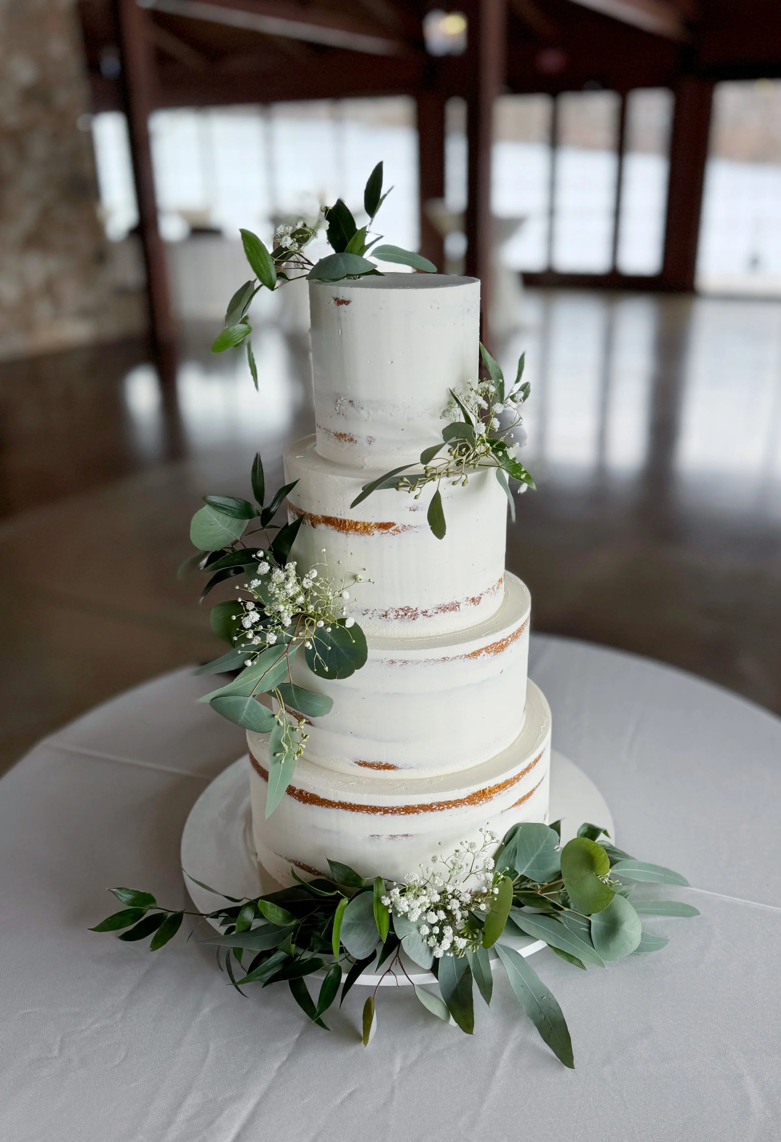Four-tier wedding cake with semi-naked frosting, decorated with greenery and white flowers, displayed on a white tablecloth.