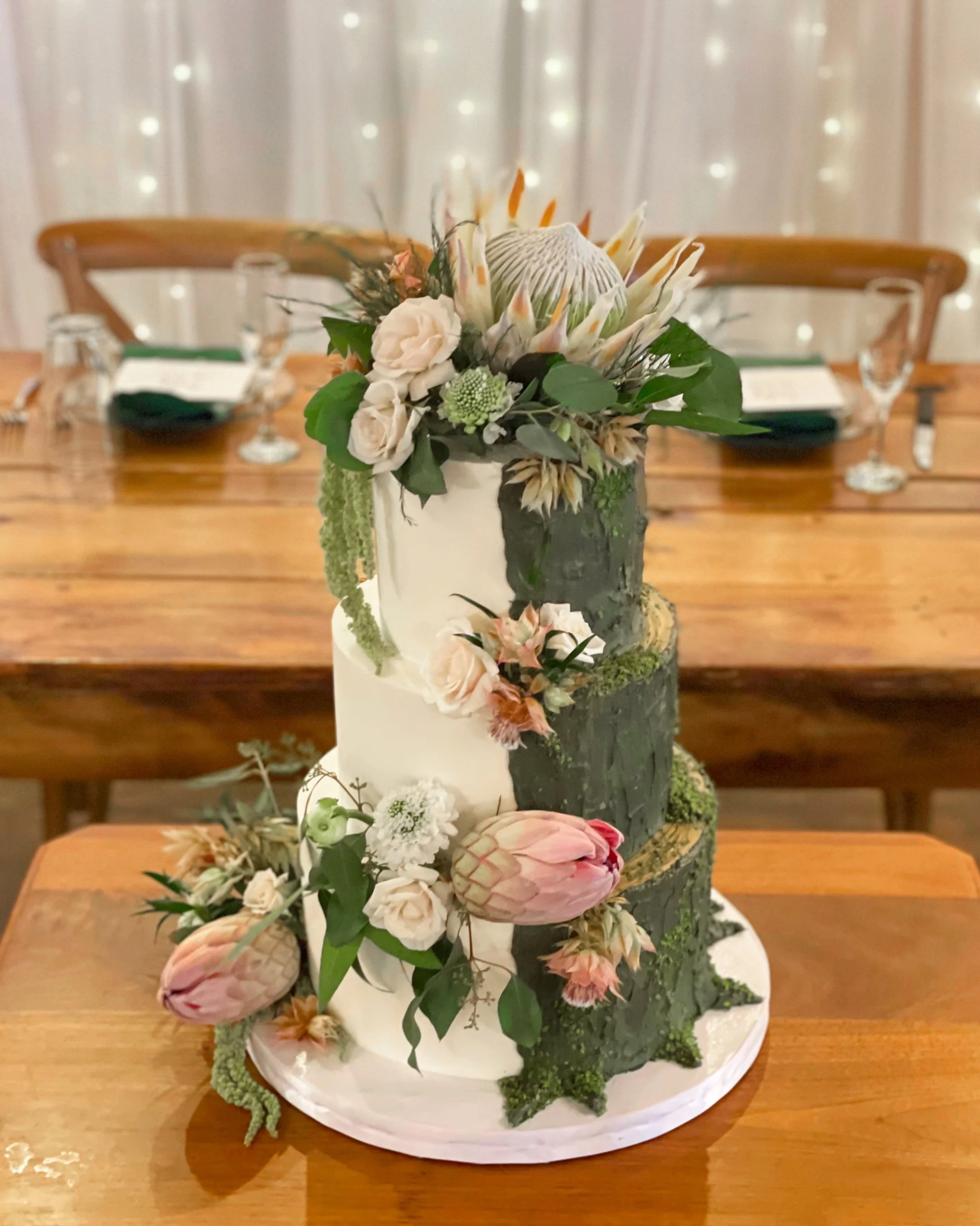 A three-tier wedding cake with a woodland theme, decorated with fresh flowers and greenery, placed on a wooden table.