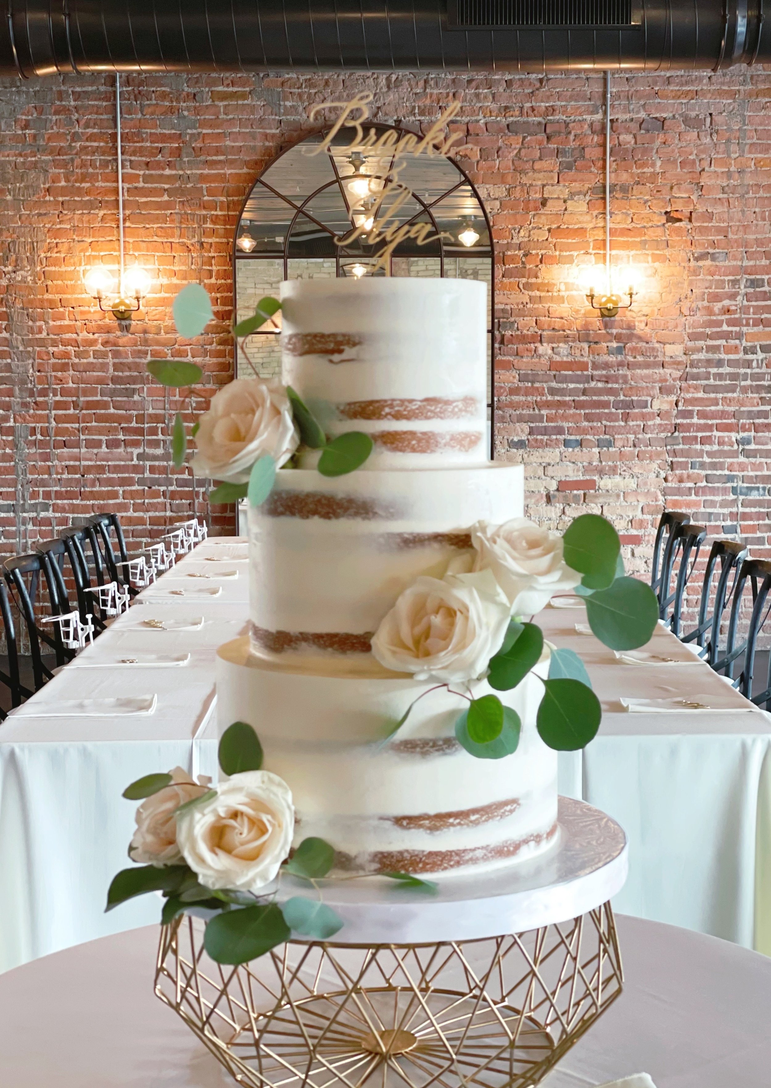 Three-tier wedding cake with white frosting, decorated with white roses and green leaves, placed on a gold geometric cake stand in a decorated reception area with brick walls and a mirror.