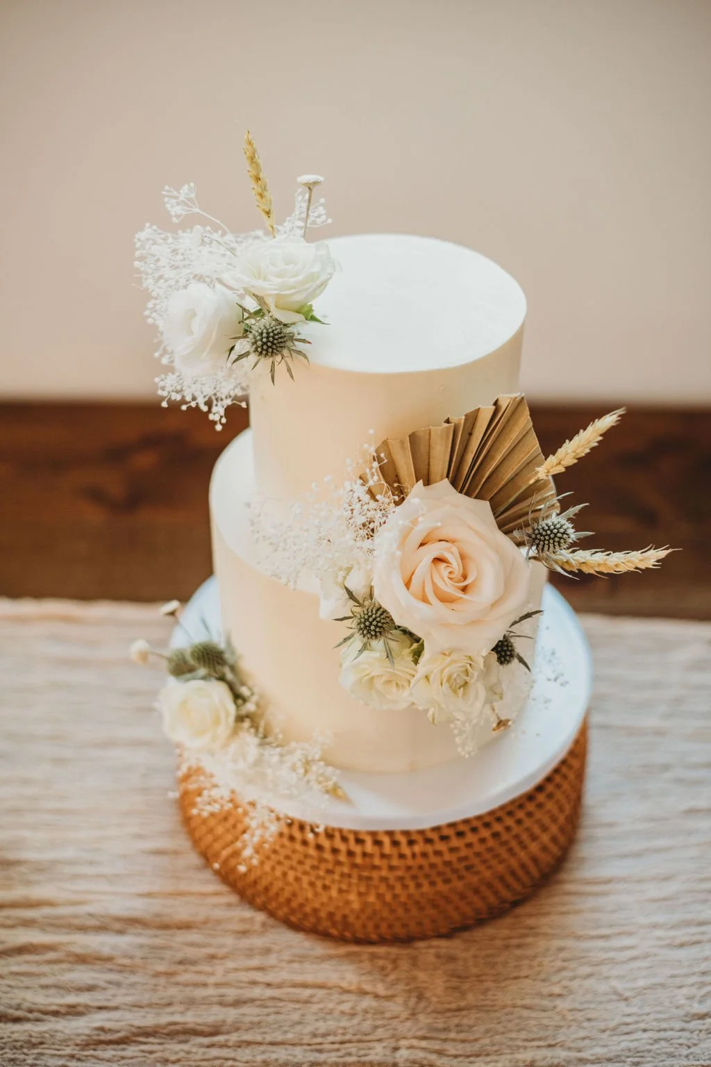 A two-tiered white wedding cake decorated with white and blush roses, dried flowers, and wheat, with a textured gold base, on a wooden surface.
