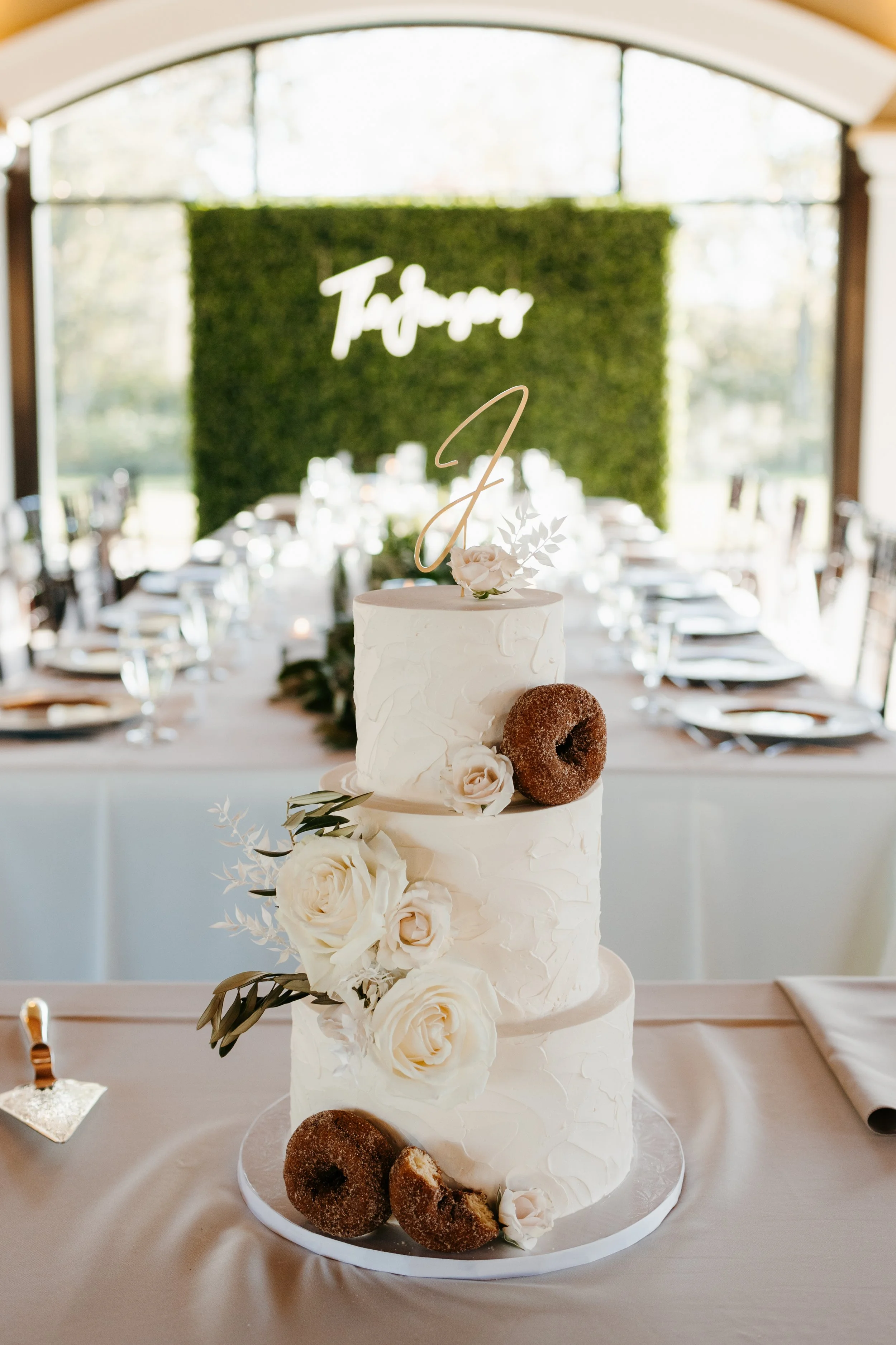 A three-tier white wedding cake decorated with white roses and greenery, topped with a gold 'J' cake topper, accompanied by donuts on the table, with a decorated dining area in the background.