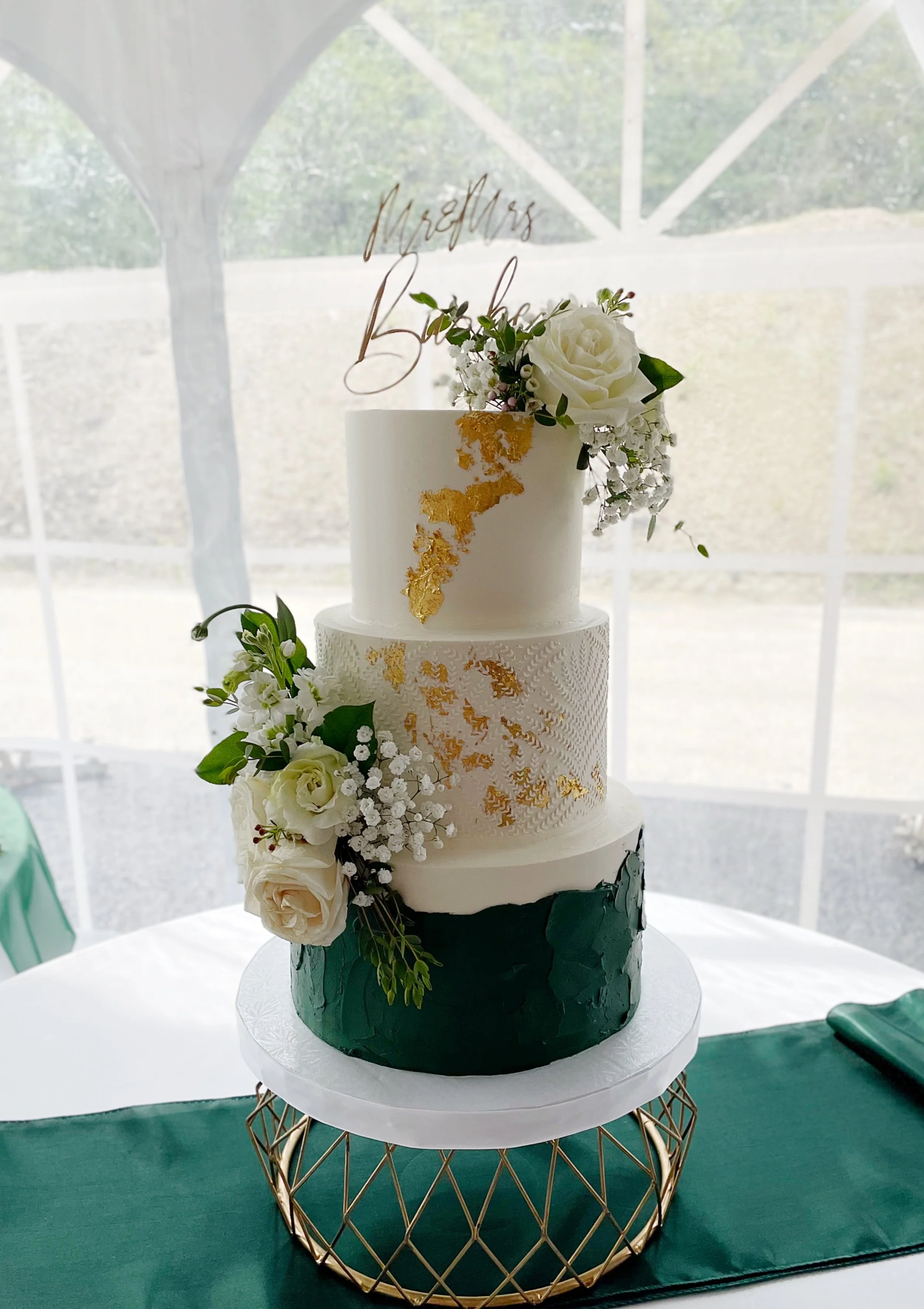 Three-tiered wedding cake with gold accents and white flowers on top and side, sitting on a decorative gold stand.