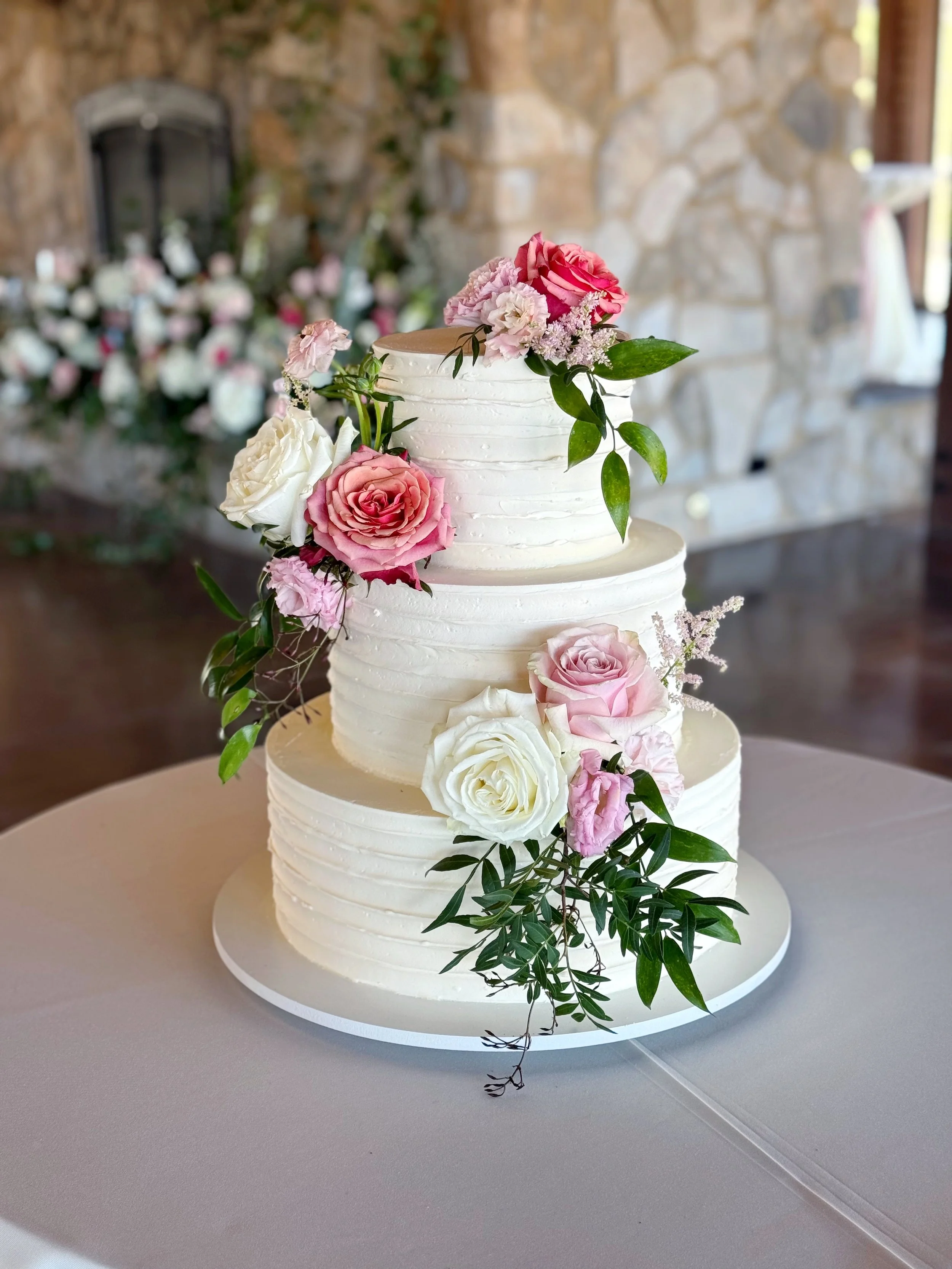 A three-tier white wedding cake decorated with pink and white roses and greenery on a white tablecloth.