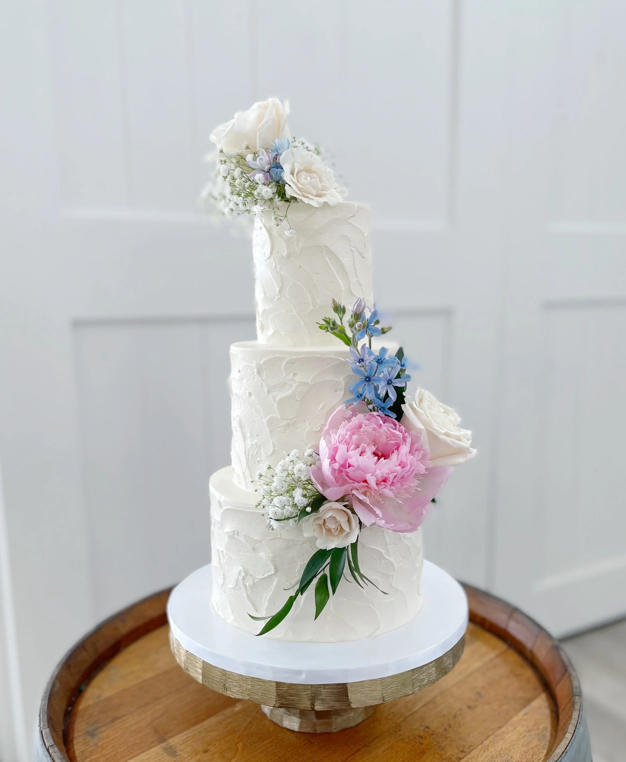 A four-tier white wedding cake decorated with fresh pink, white, and blue flowers and greenery, placed on a round wooden table.