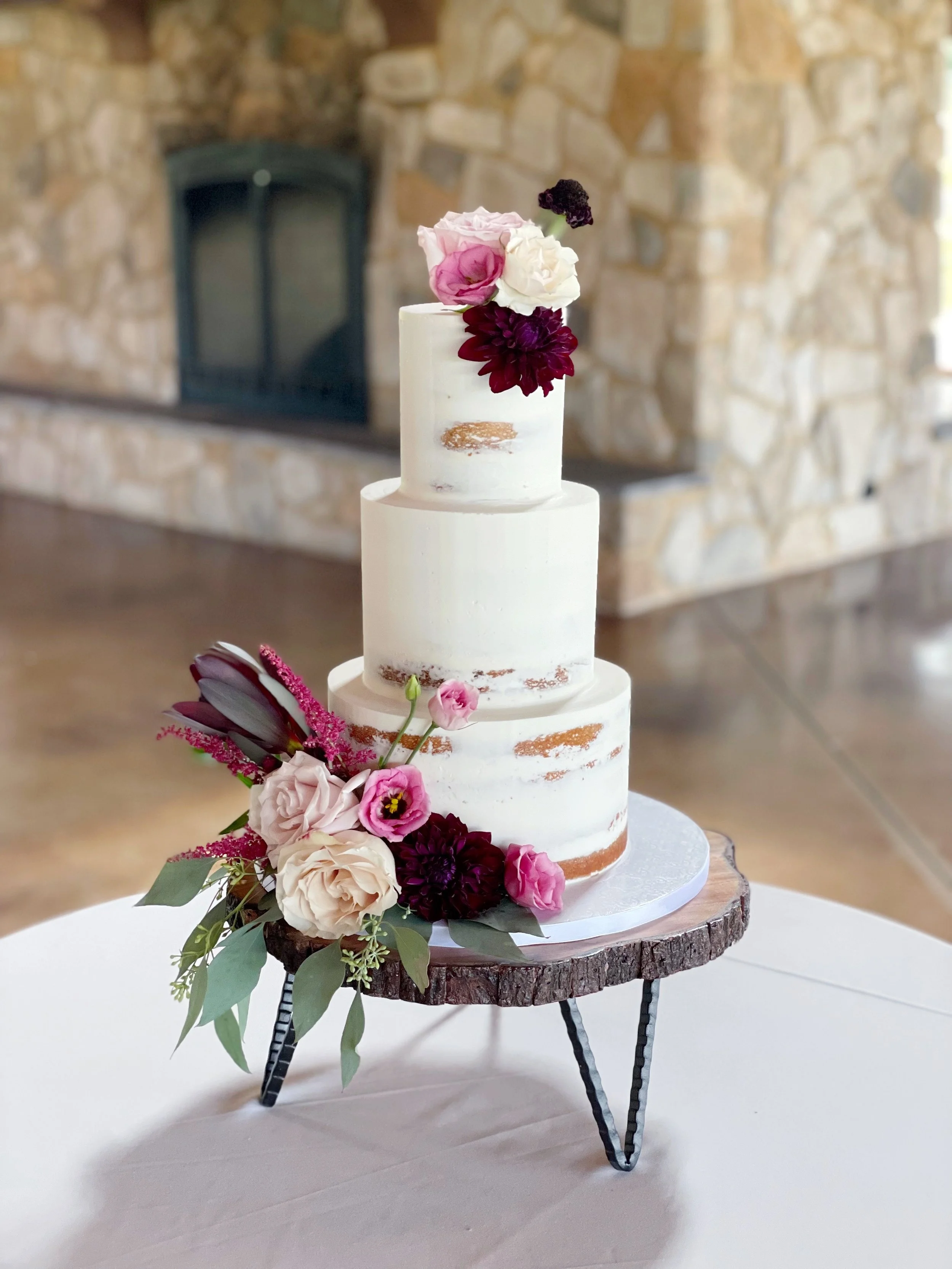Three-tiered white wedding cake decorated with pink, white, and dark purple flowers, placed on a wooden cake stand on a white table.