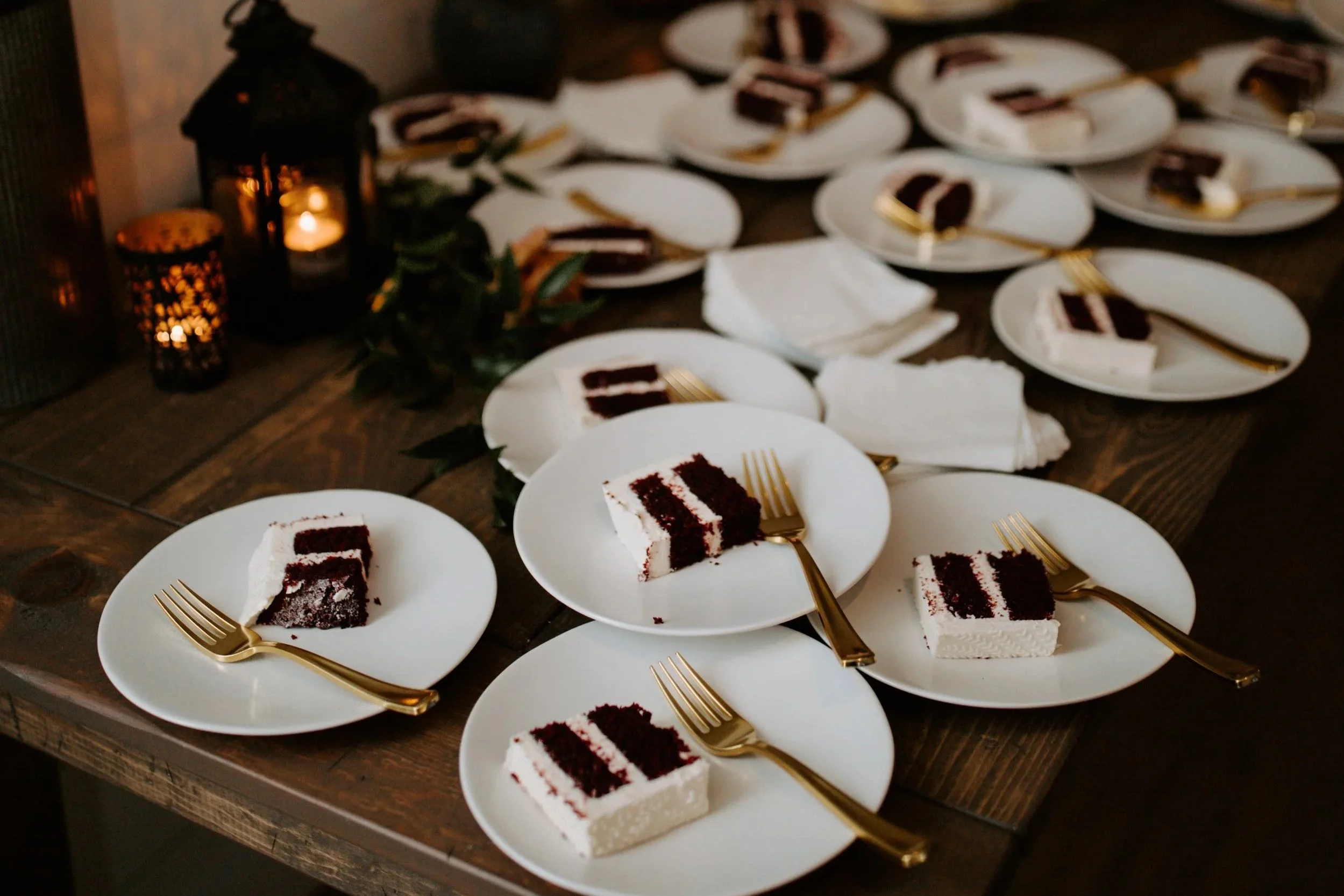 Multiple plates with slices of chocolate cake and gold forks on a wooden table, decorated with candle lanterns and greenery.
