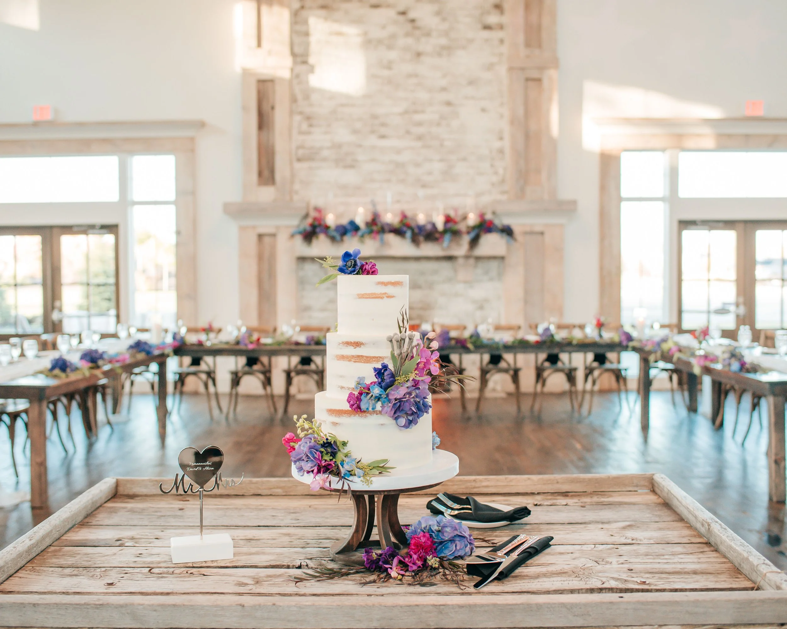 A wedding cake decorated with purple, pink, and blue flowers placed on a wooden table, with a small sign reading 'Mr. & Mrs.' nearby. The background shows a reception hall with long tables, floral decorations, large windows, and a stone fireplace.