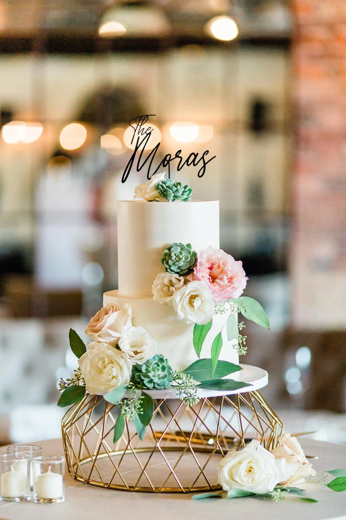 A two-tiered white wedding cake adorned with pink and white roses, succulents, and green leaves, with a black cake topper that reads 'The Moras'. The cake is on a decorative gold stand, and there are small candles and flowers on the table.
