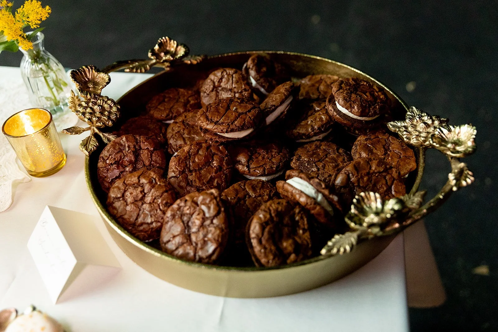 A gold decorative tray filled with cookies, a small yellow candle holder, and a glass vase with yellow flowers on a white tablecloth.