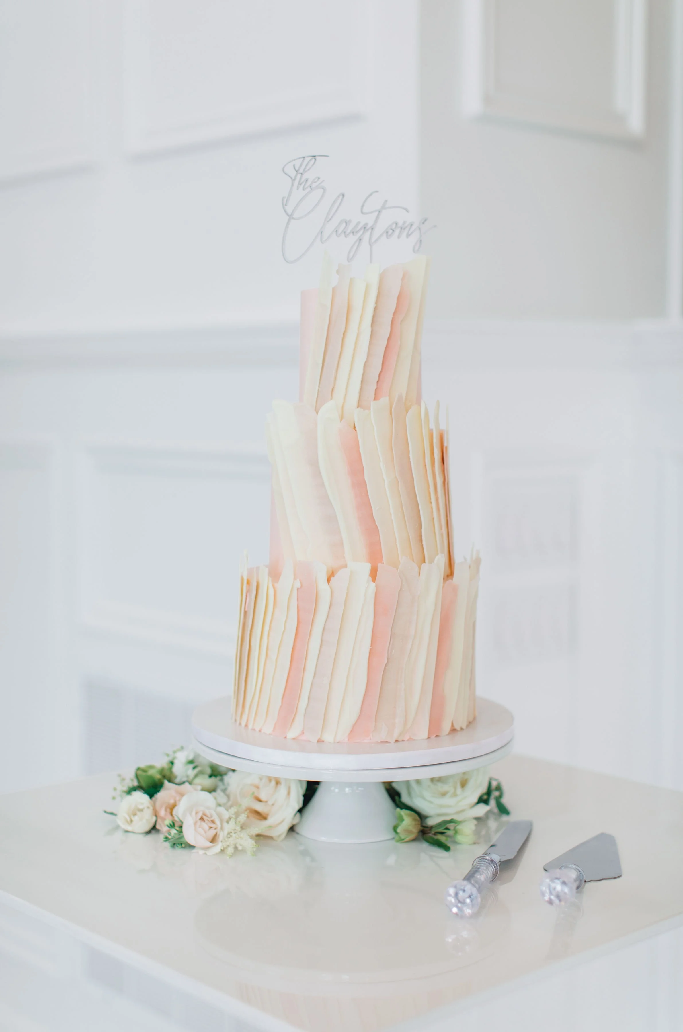 A three-tier wedding cake decorated with pale pink and white ruffle-like fondant strips, topped with a clear acrylic cake topper that reads 'The Clayton's', surrounded by a small floral arrangement with pale pink roses and greenery, with cake knives 