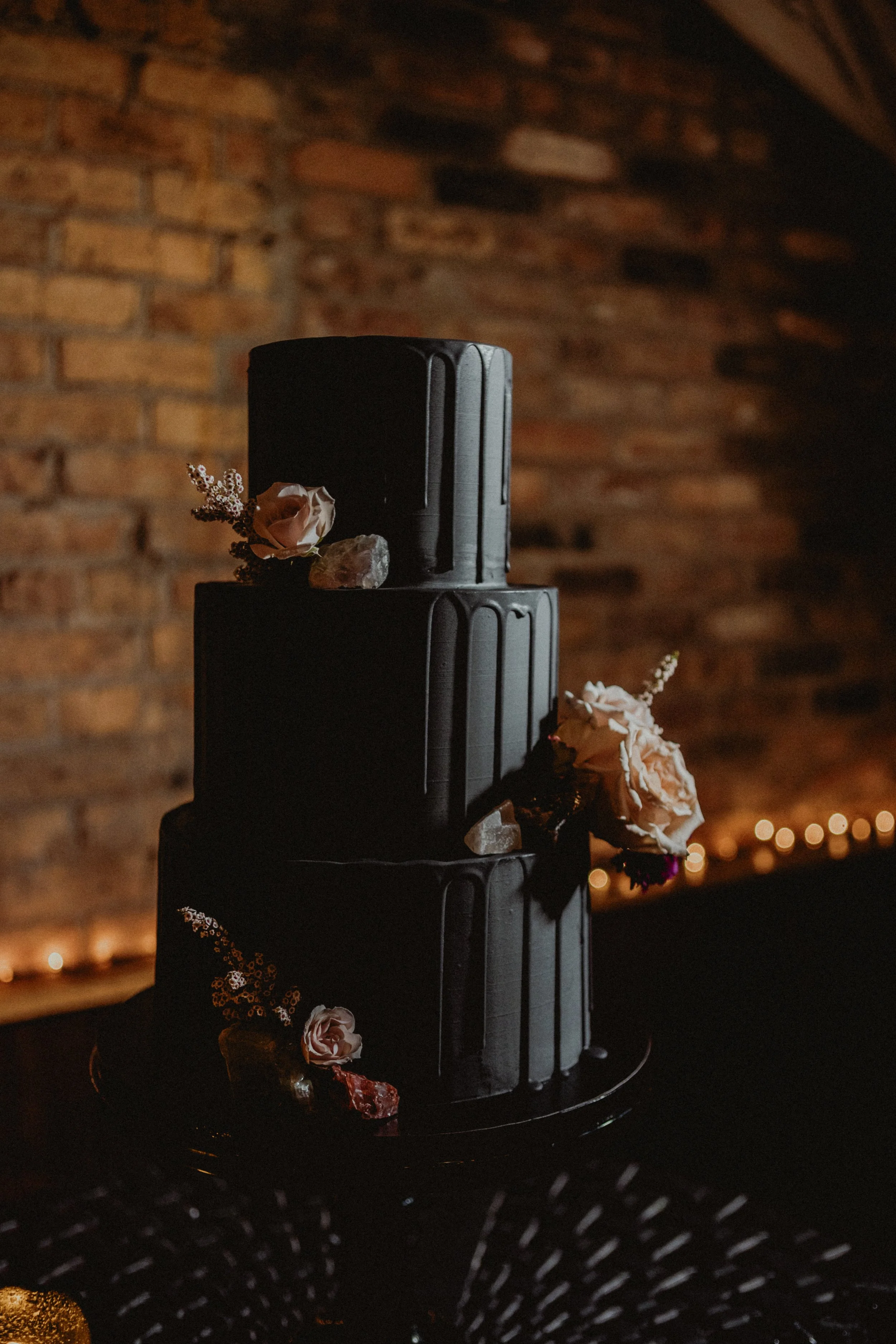 Three-tier black wedding cake decorated with flowers, crystals, and stones, against a brick wall background.
