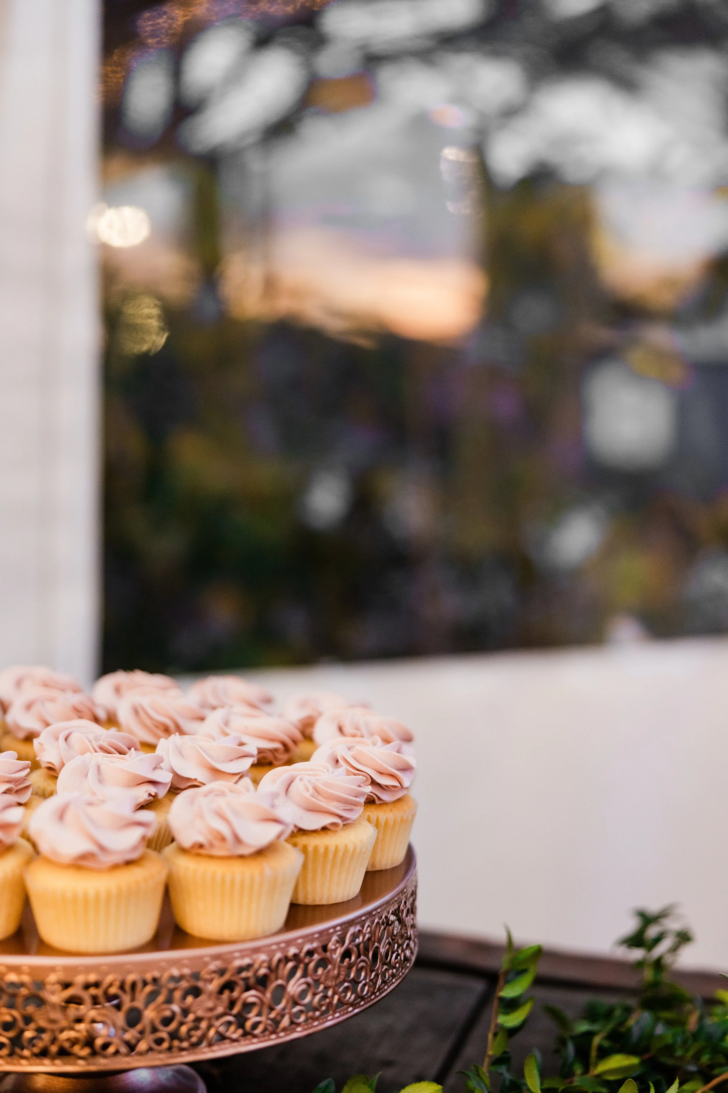 Pink frosted cupcakes arranged on a decorative rose gold cake stand with a blurred window and sunset in the background.