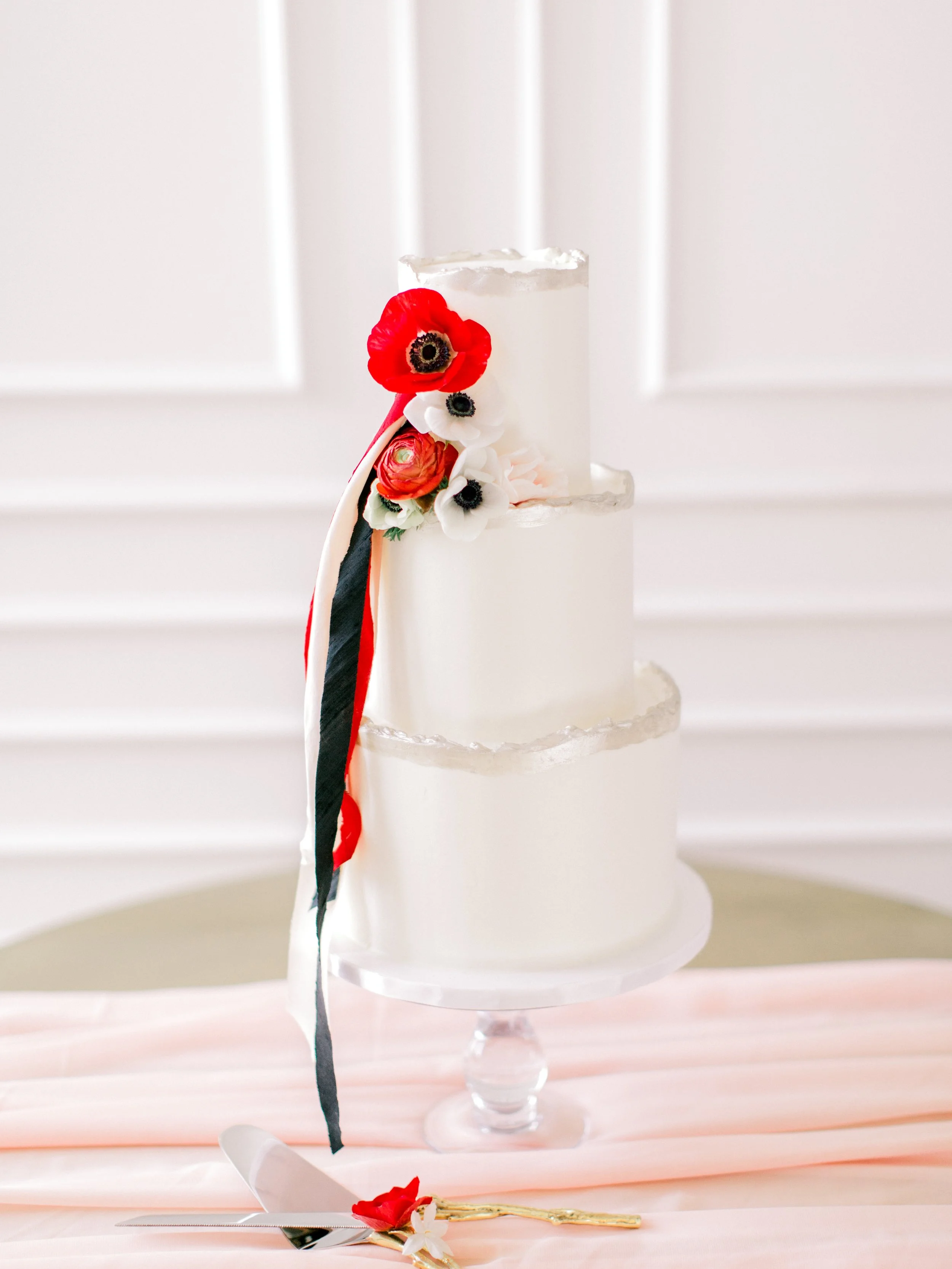 Three-tiered white wedding cake decorated with red, white, and black flowers and ribbons, set on a glass cake stand.