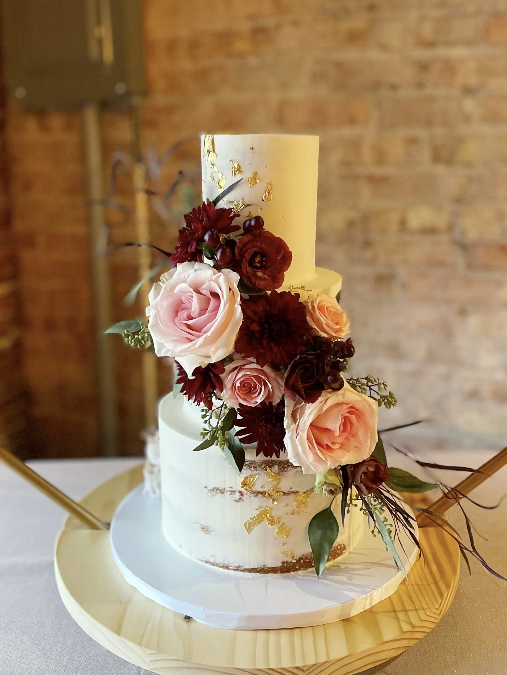 A two-tiered wedding cake decorated with fresh pink, peach, and burgundy flowers and gold leaf accents, displayed on a wooden cake stand.