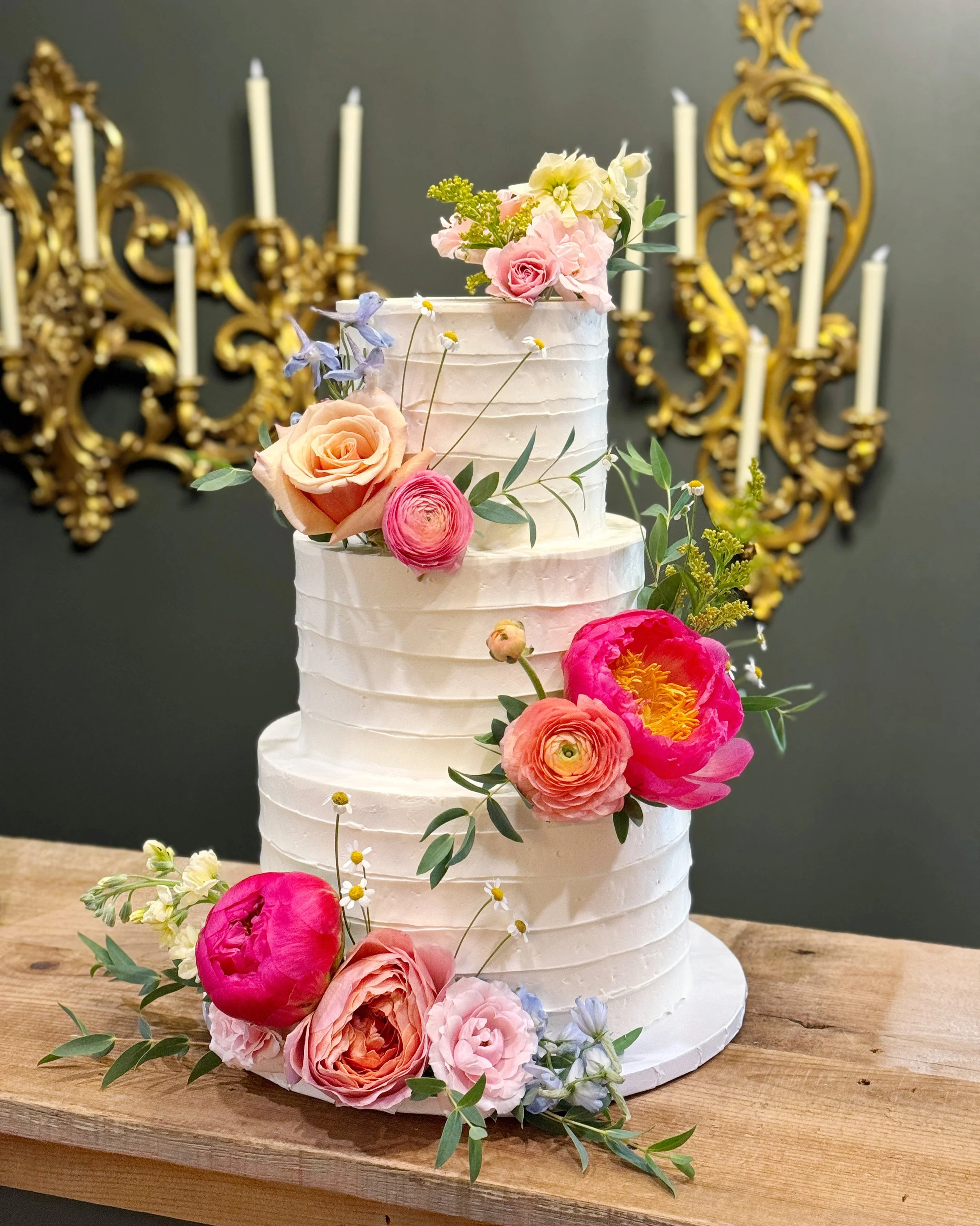 Three-tier white wedding cake decorated with pink, peach, and purple flowers and green foliage, placed on a wooden table in front of gold ornamental wall decor and white candlesticks.