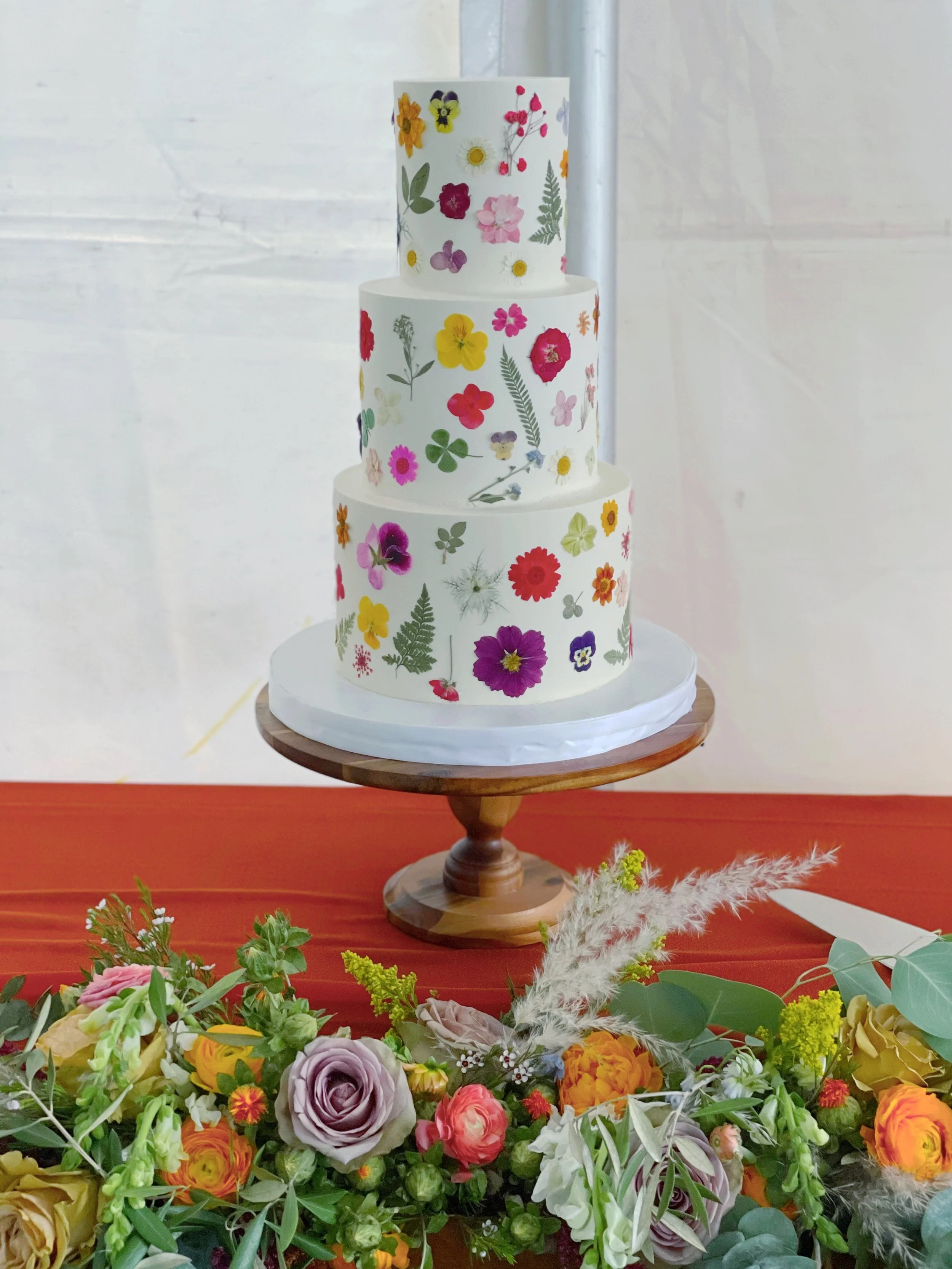 Three-tiered white wedding cake decorated with colorful floral patterns, placed on a wooden cake stand on a red table, with a flower arrangement in front.